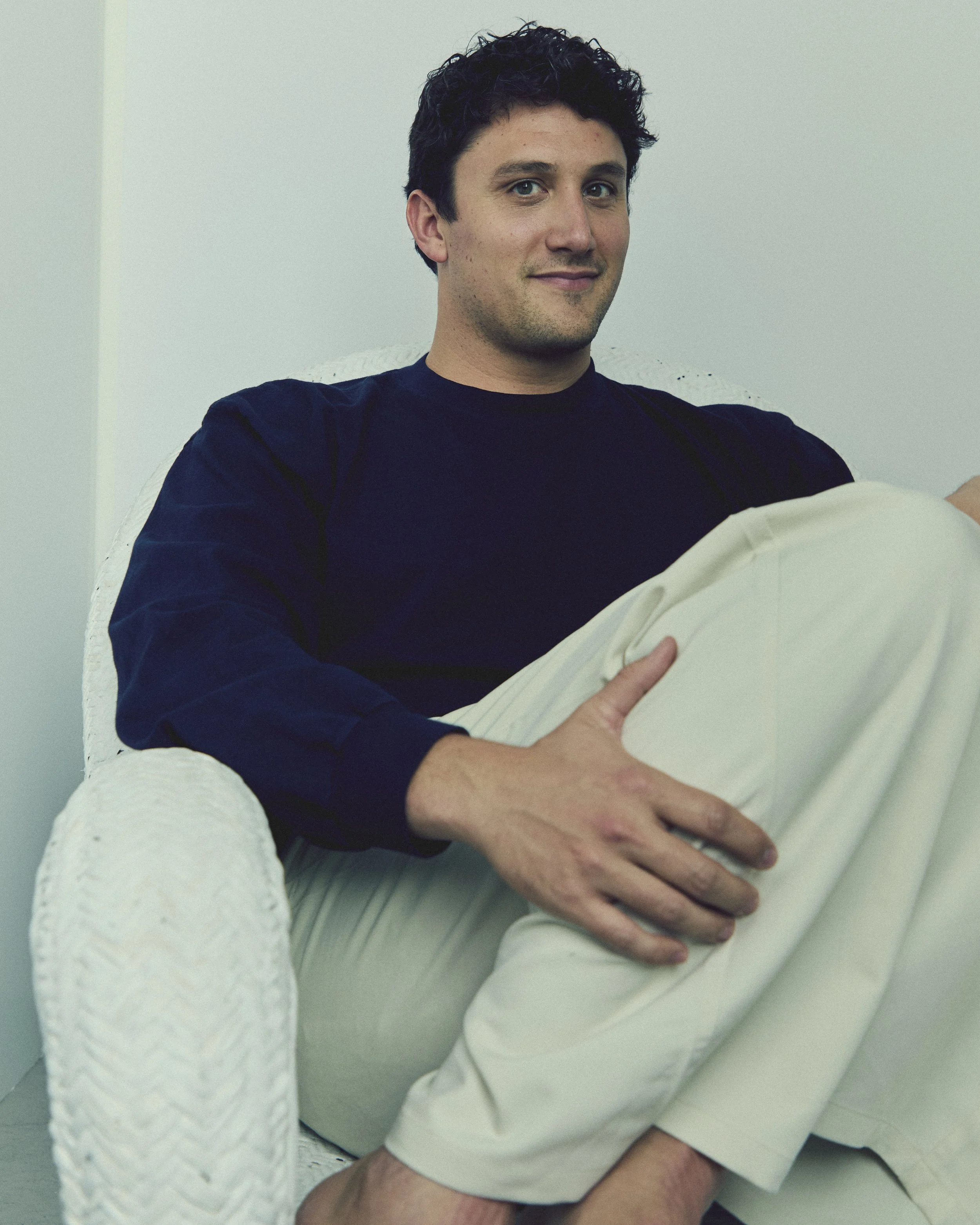 A man with dark curly hair sitting on a white wicker chair, wearing a black long sleeve shirt and beige pants, with a relaxed and slight smile.