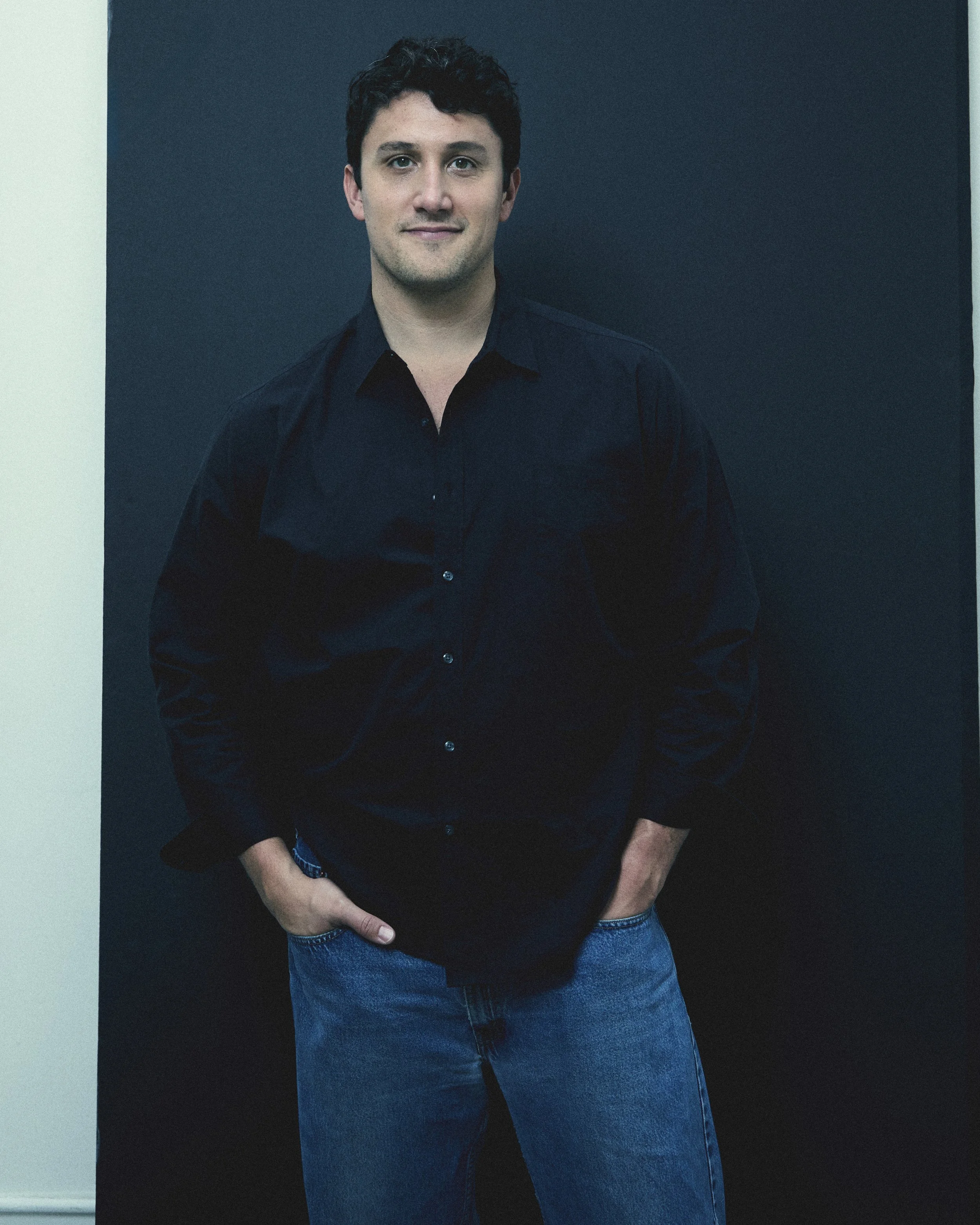 A young man with dark curly hair wearing a black shirt and blue jeans, standing against a black background with a slight smile.