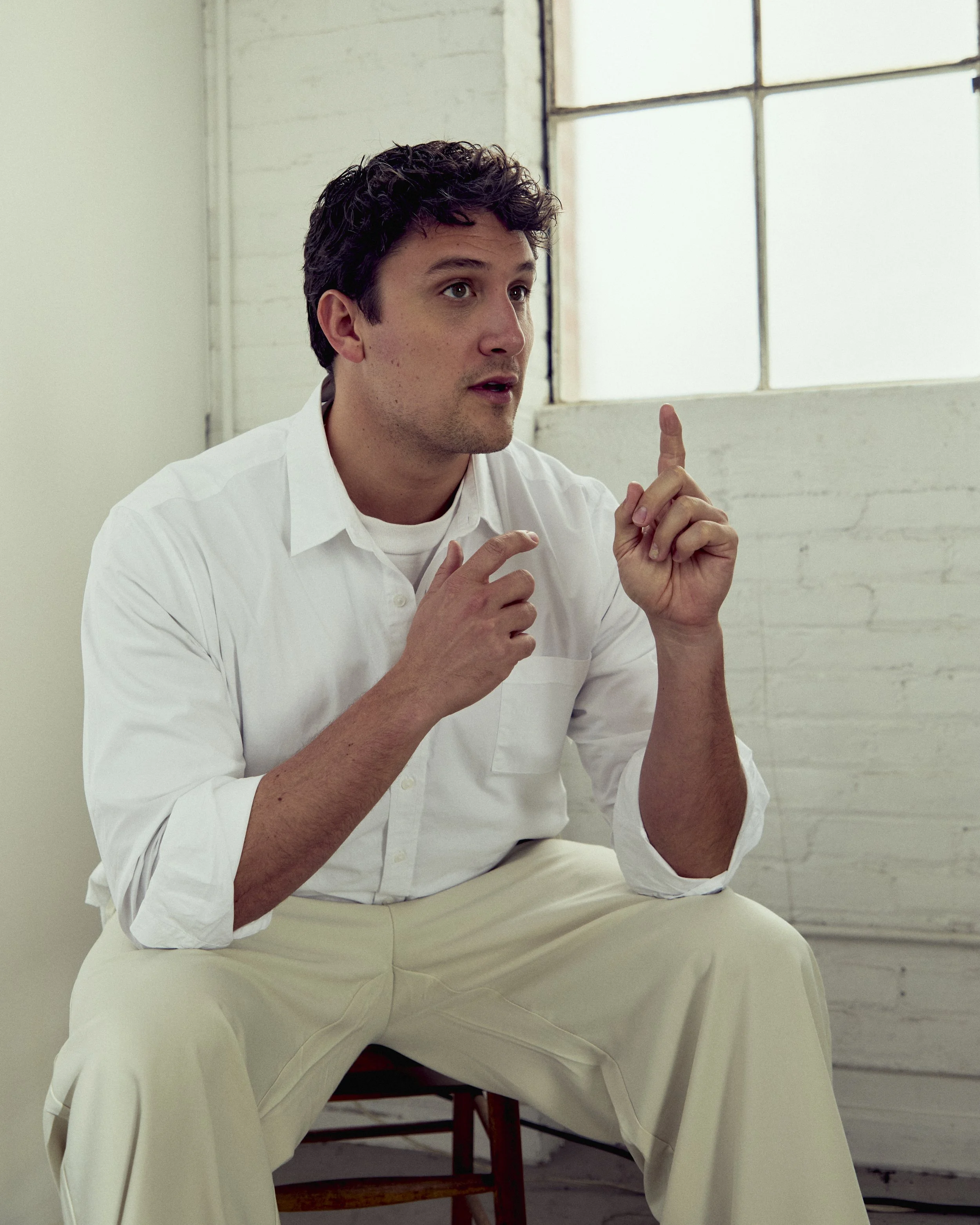 A man in a white shirt and beige pants sitting on a wooden chair, gesturing with his right hand and holding his left hand near his face, inside a room with a large window in the background.