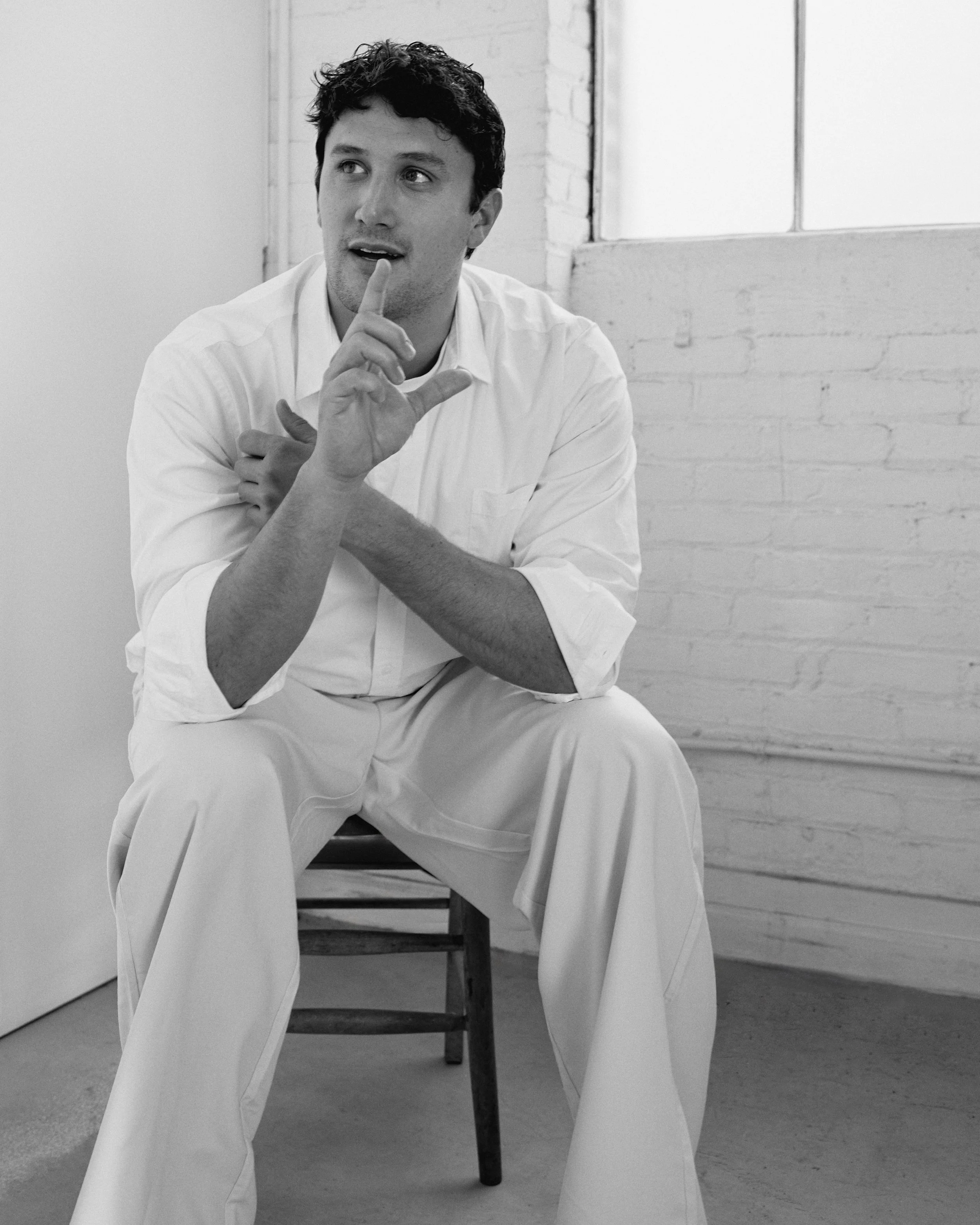 A man with dark, curly hair sitting on a wooden chair in a room with a white brick wall and a window. He is wearing a white shirt and white pants, with a thoughtful expression, holding one hand up with a finger to his lips.