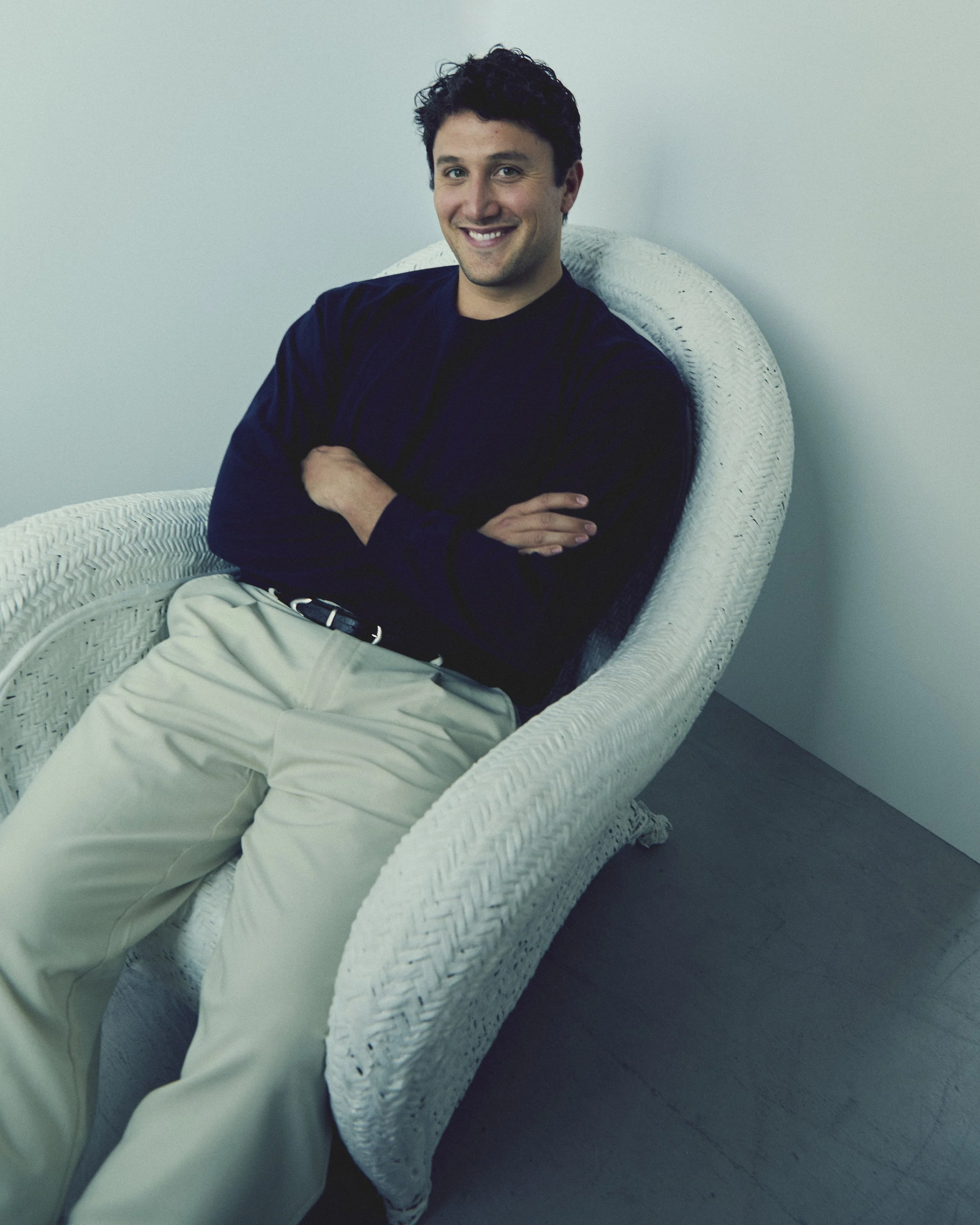 A man sitting in a white wicker chair, smiling, with arms crossed, wearing a black shirt and beige pants, against a plain light-colored wall.