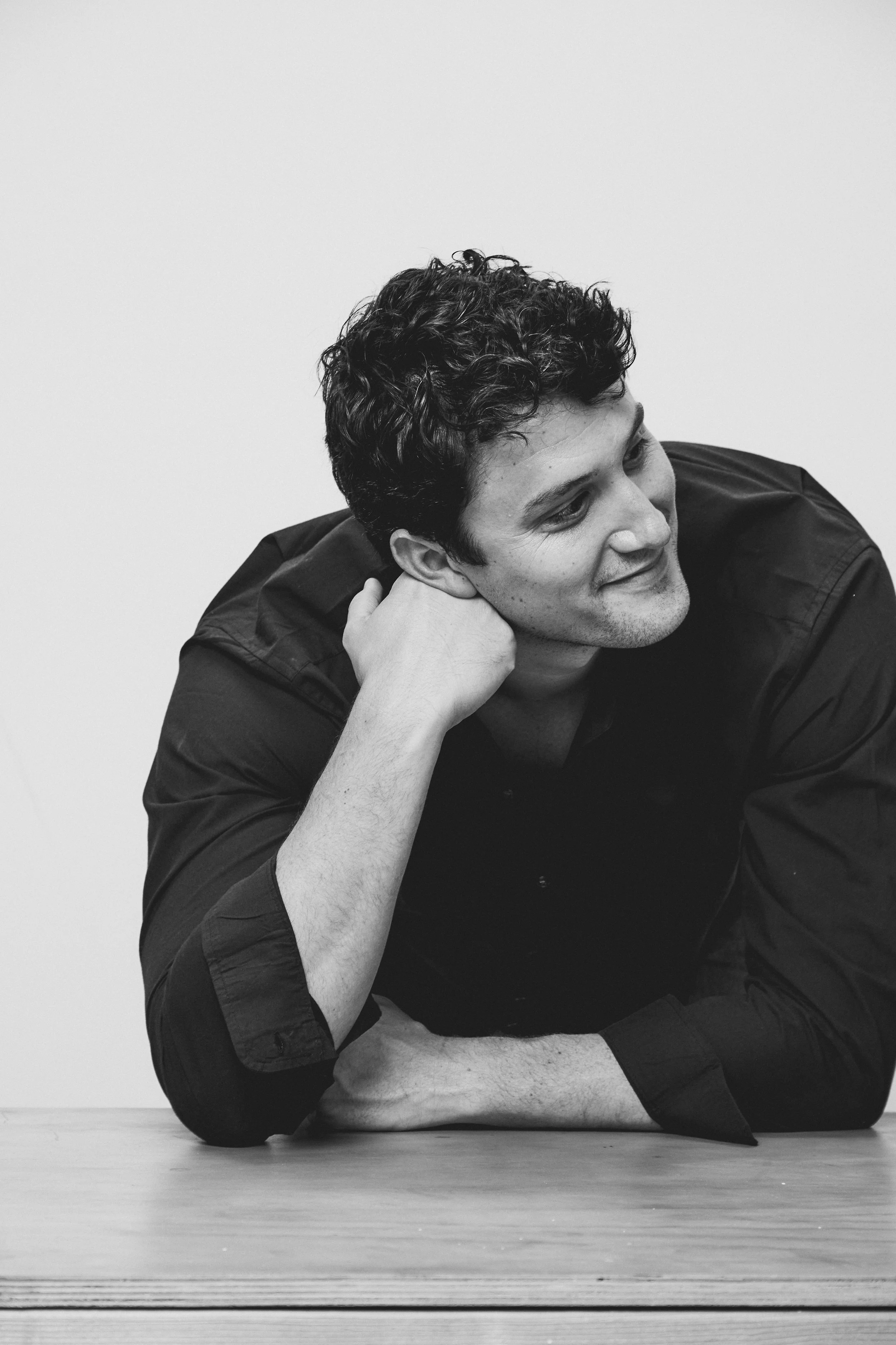 A young man with curly hair, wearing a dark shirt, is resting his head on his hand, leaning on a wooden surface, and smiling softly while looking to the side against a plain background.
