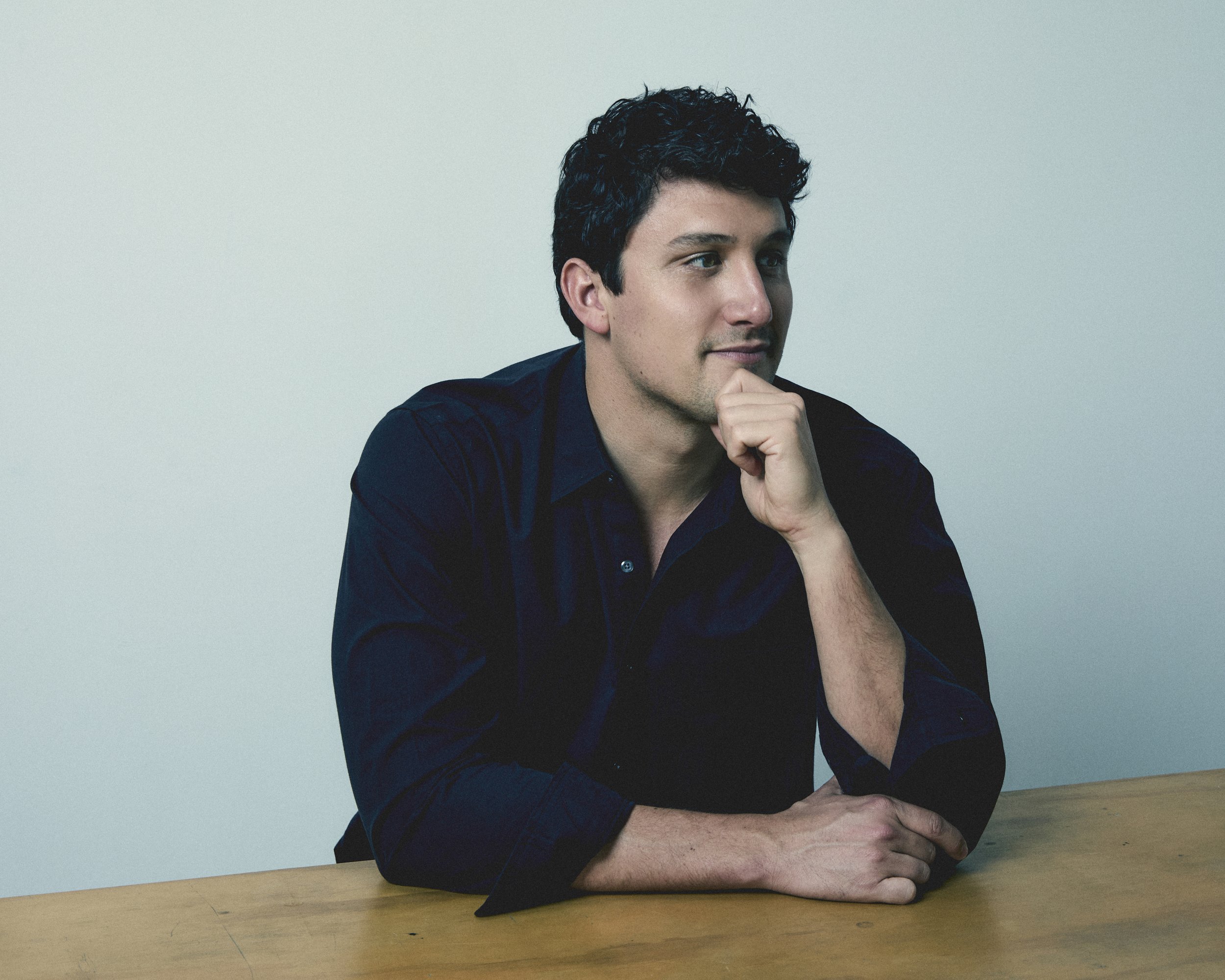 A young man with dark hair and wearing a dark shirt, sitting at a wooden table with his chin resting on his hand, looking thoughtfully to the side
