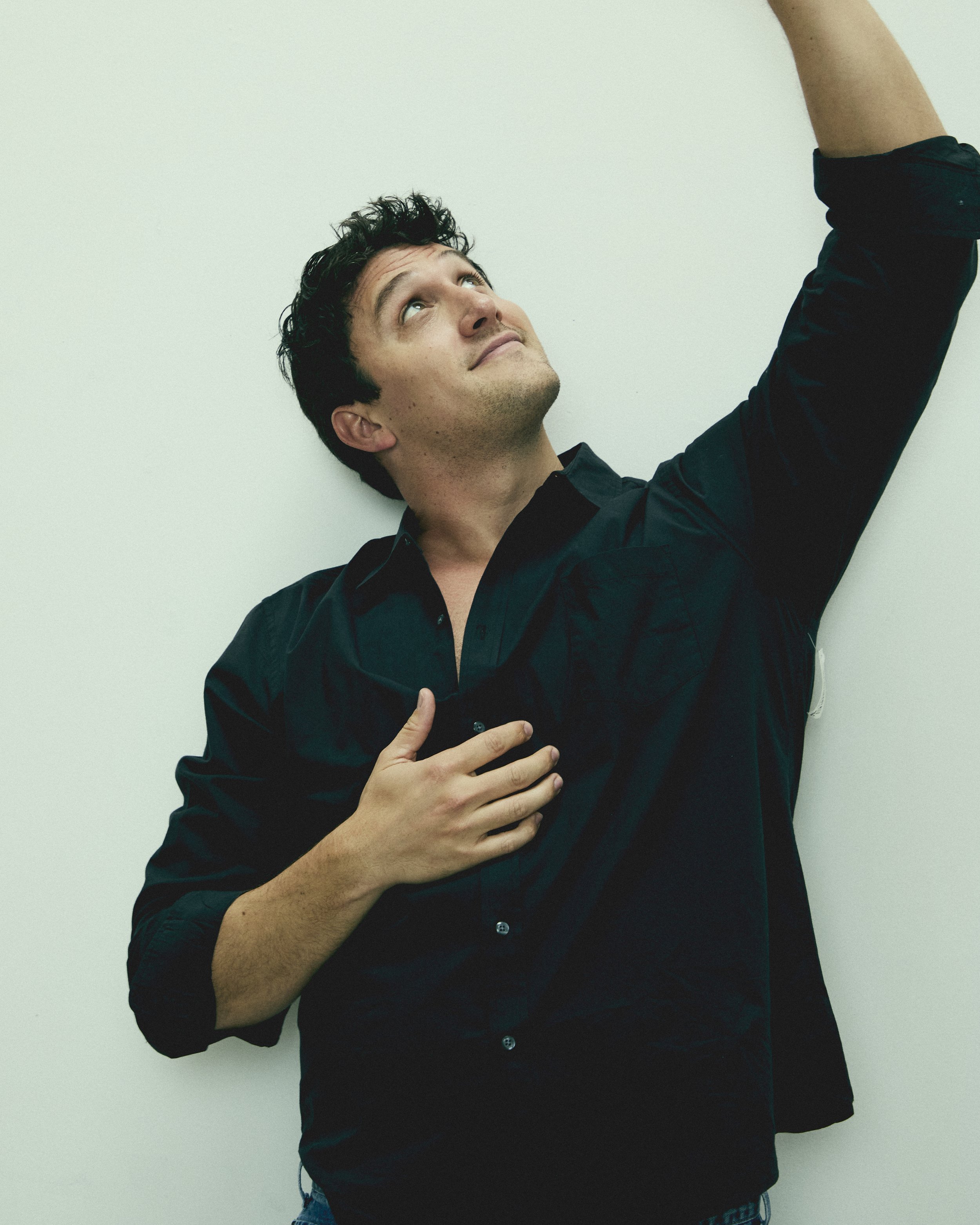 A man with dark curly hair and a black shirt stands against a plain light-colored wall. He is looking upward, with one hand on his chest and the other arm raised above his head.