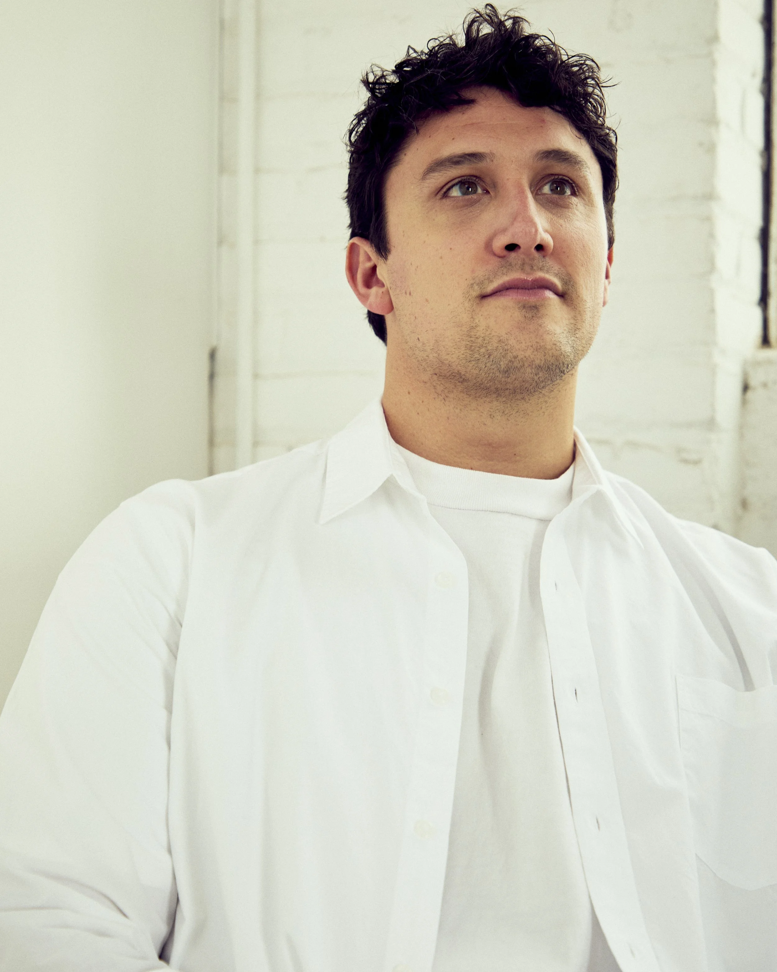 Young man with dark curly hair wearing a white shirt looking slightly upward in a room with white brick walls.