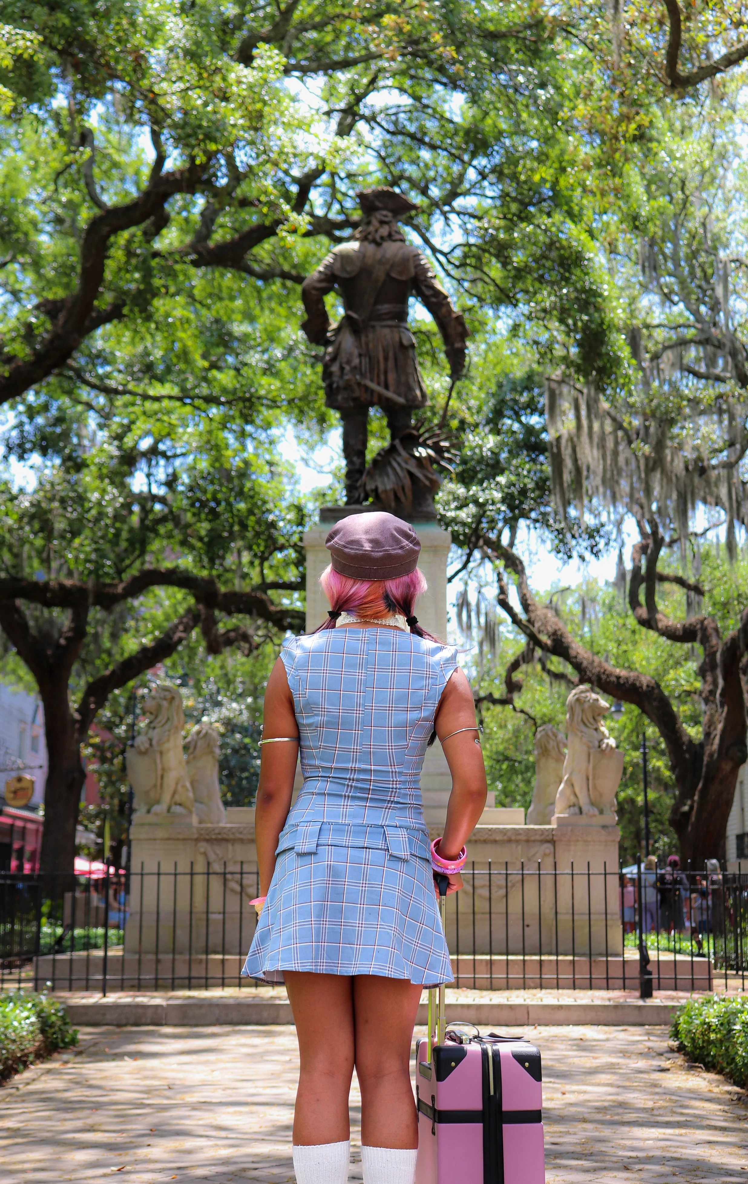 A woman with pink hair wearing a plaid dress, standing with her back to the camera, looking at a statue of a man in historical clothing on a pedestal amidst trees and lion sculptures.