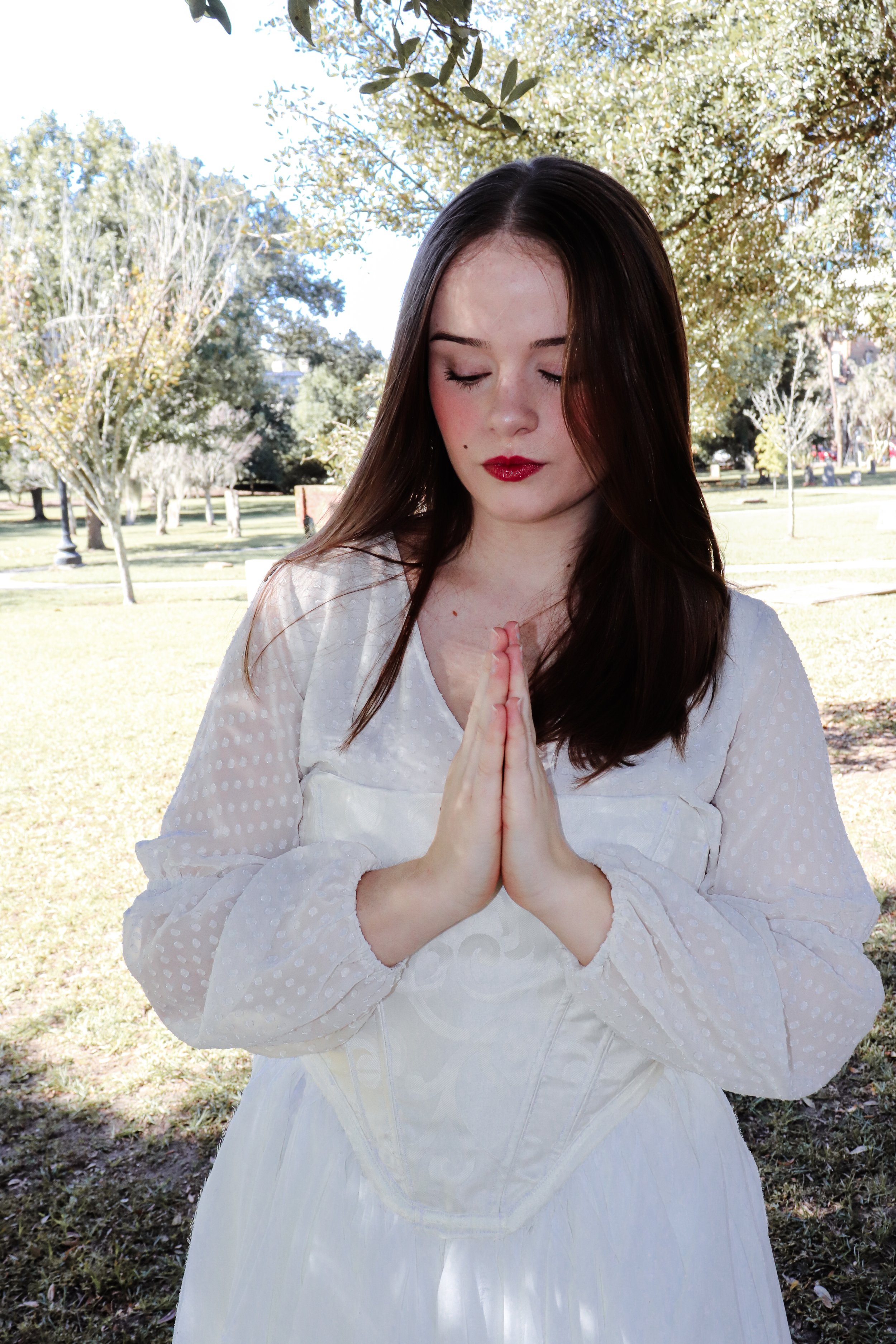 A young woman with long brown hair and fair skin, wearing a white dress, stands outdoors with her eyes closed and hands pressed together in a prayer-like gesture. She is in a park with trees and grass visible in the background on a sunny day.