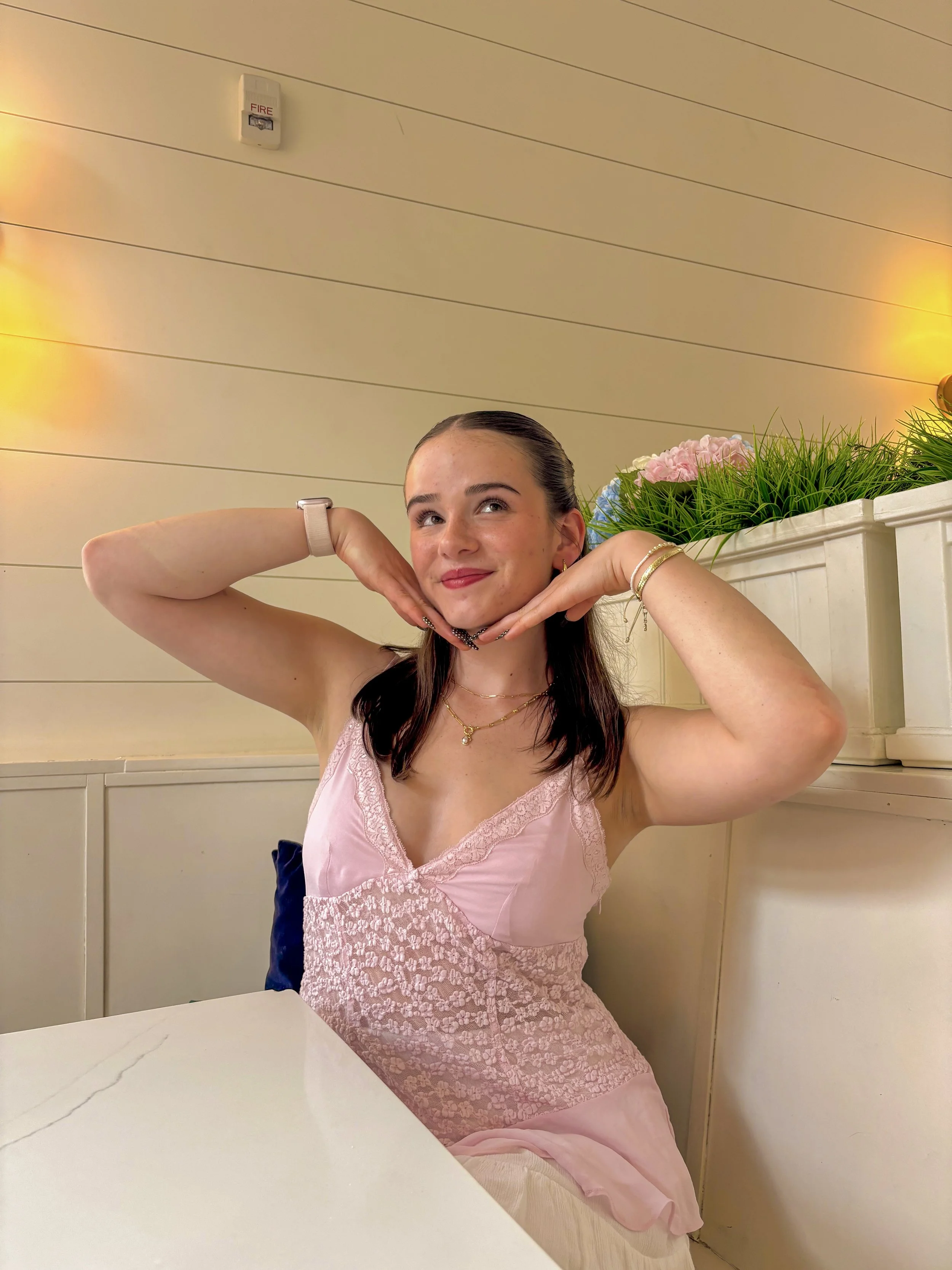 A young woman with dark brown hair, wearing a pink lace top, sitting at a table in a restaurant, resting her chin on her hands with a playful smile. There are pink roses and greenery on a ledge behind her, and warm yellow lighting on the wall.