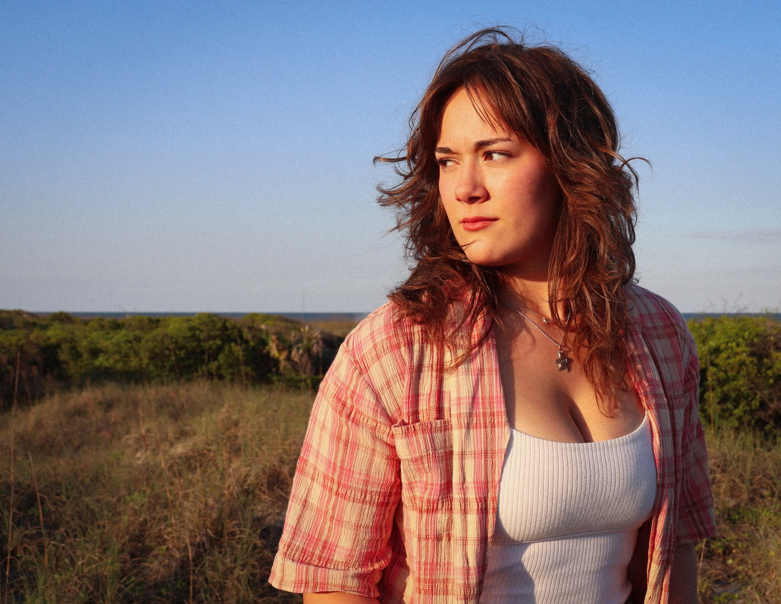 A woman with wavy brown hair wearing a white tank top and a red plaid shirt standing outdoors on a grassy field during sunset, looking to her right.
