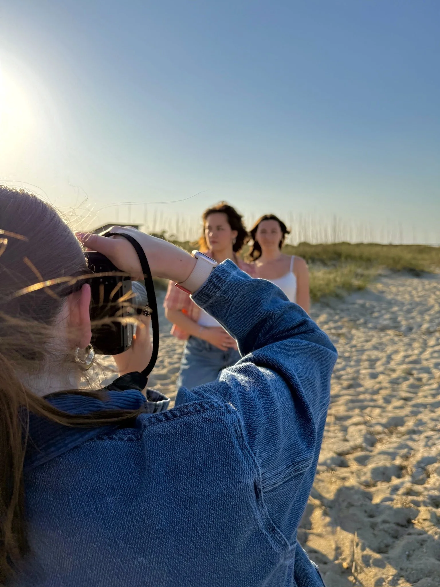 A person is taking a photo of two women standing on a sandy beach during sunset.