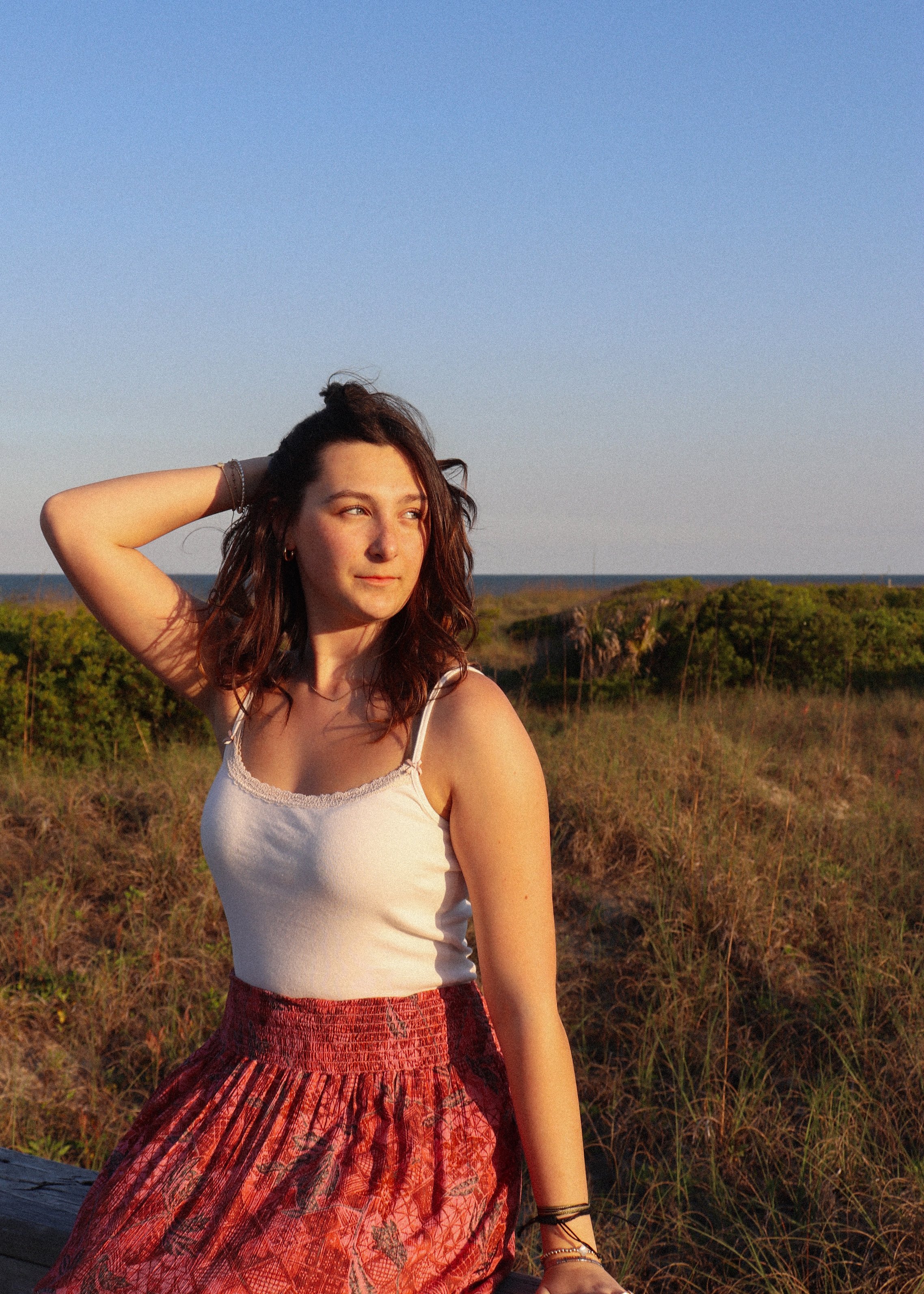 A young woman with brunette hair and light skin standing outdoors during sunset, wearing a white tank top and a red patterned skirt, looking to the side with one hand resting on her head, and a landscape with grass and bushes behind her.