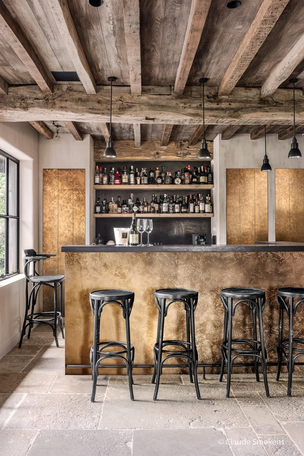 Un bar intérieur avec un comptoir en or, des tabourets noirs, des bouteilles d'alcool sur des étagères en arrière-plan, plafond en bois brut et une fenêtre à gauche.
