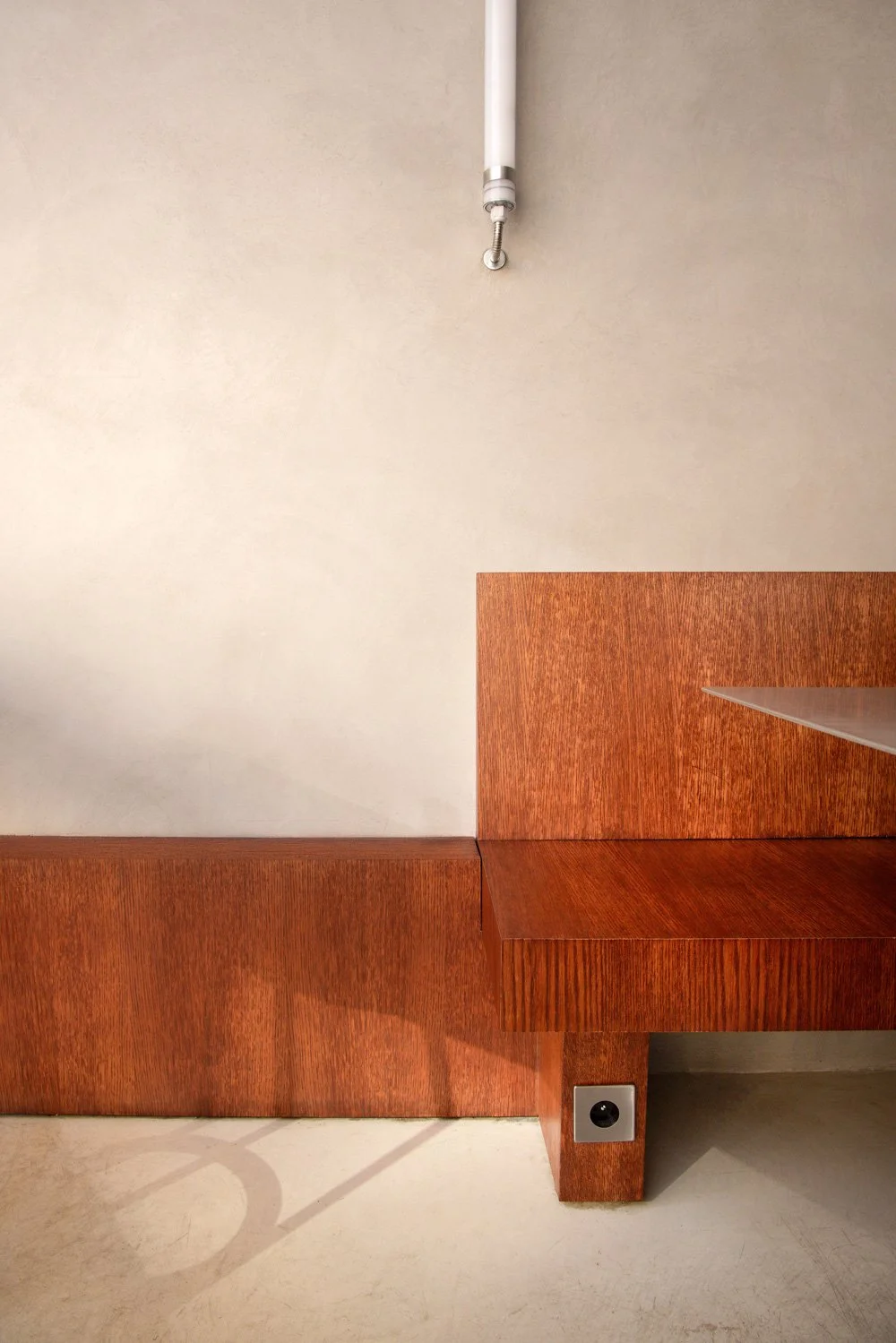 Vue d'une salle avec un mur beige et une banquette en bois rougeâtre, avec une prise électrique et une pipe d'évacuation au plafond.