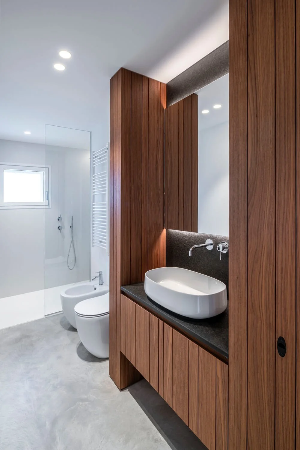 Salle de bain moderne avec meuble en bois, lavabo blanc oval, miroir, paroi en verre pour la douche, toilettes et bidet blanc, fenêtre naturelle, éclairage encastré