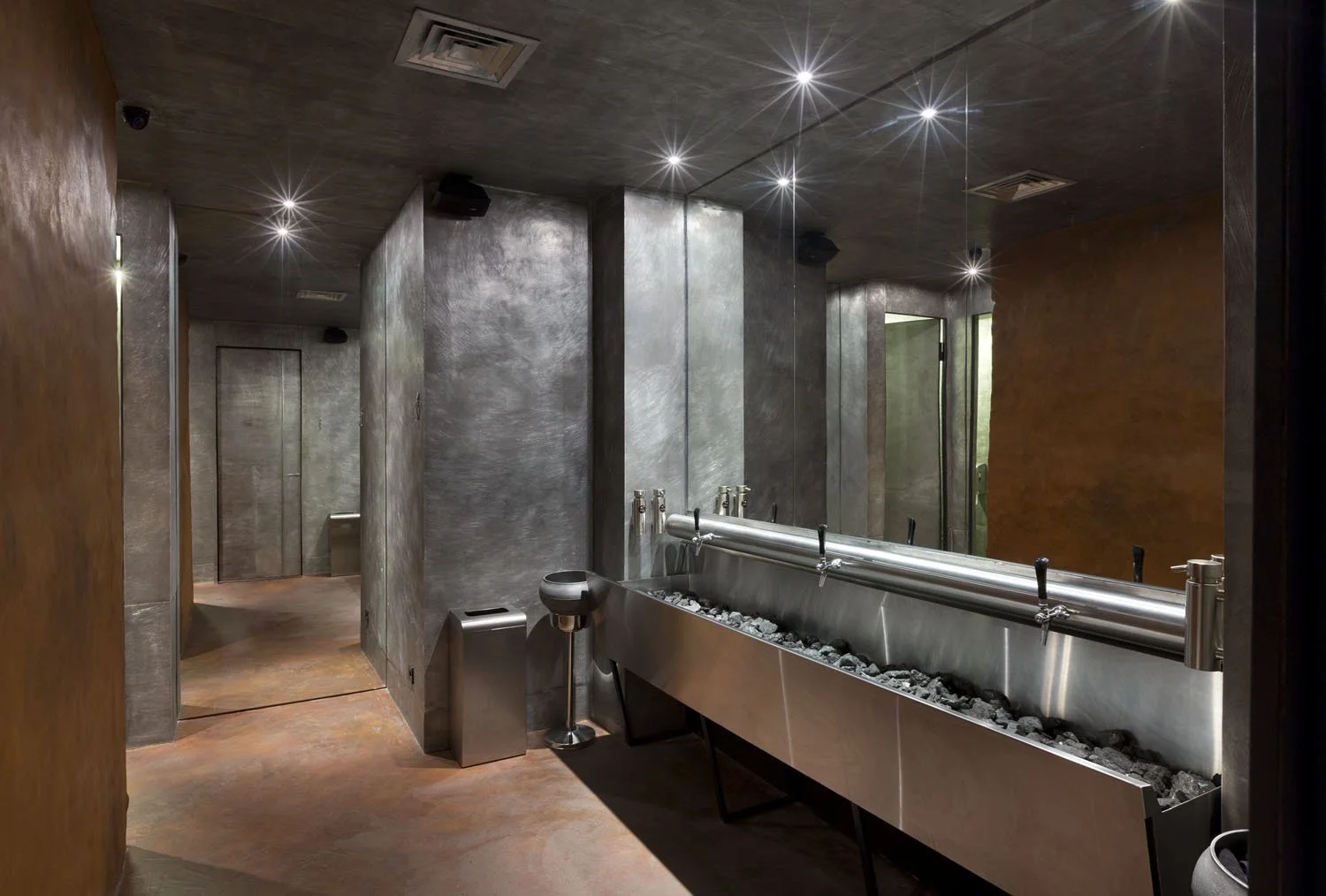Salle de relaxation ou spa avec murs en béton gris, plafond en béton, sol en terre cuite, lumière encastrée au plafond, et un banc en métal avec pierres, dans un environnement moderne et minimaliste.