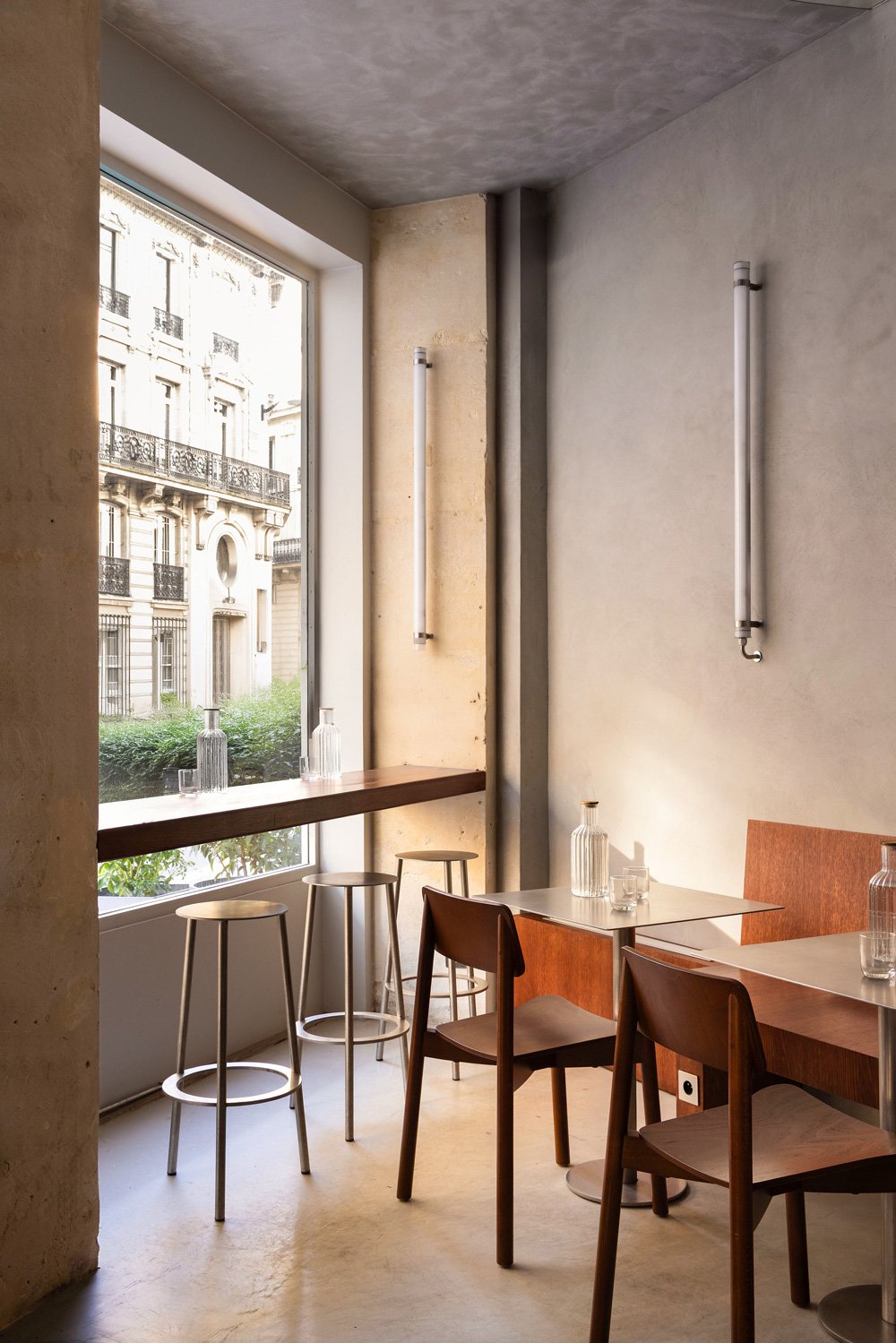Intérieur d'un café avec des tables en bois, des chaises en bois, des bouteilles en verre transparent sur les tables, un grand fenêtre laissant entrer de la lumière naturelle, et deux tubes lumineux fixés au mur gris et beige.