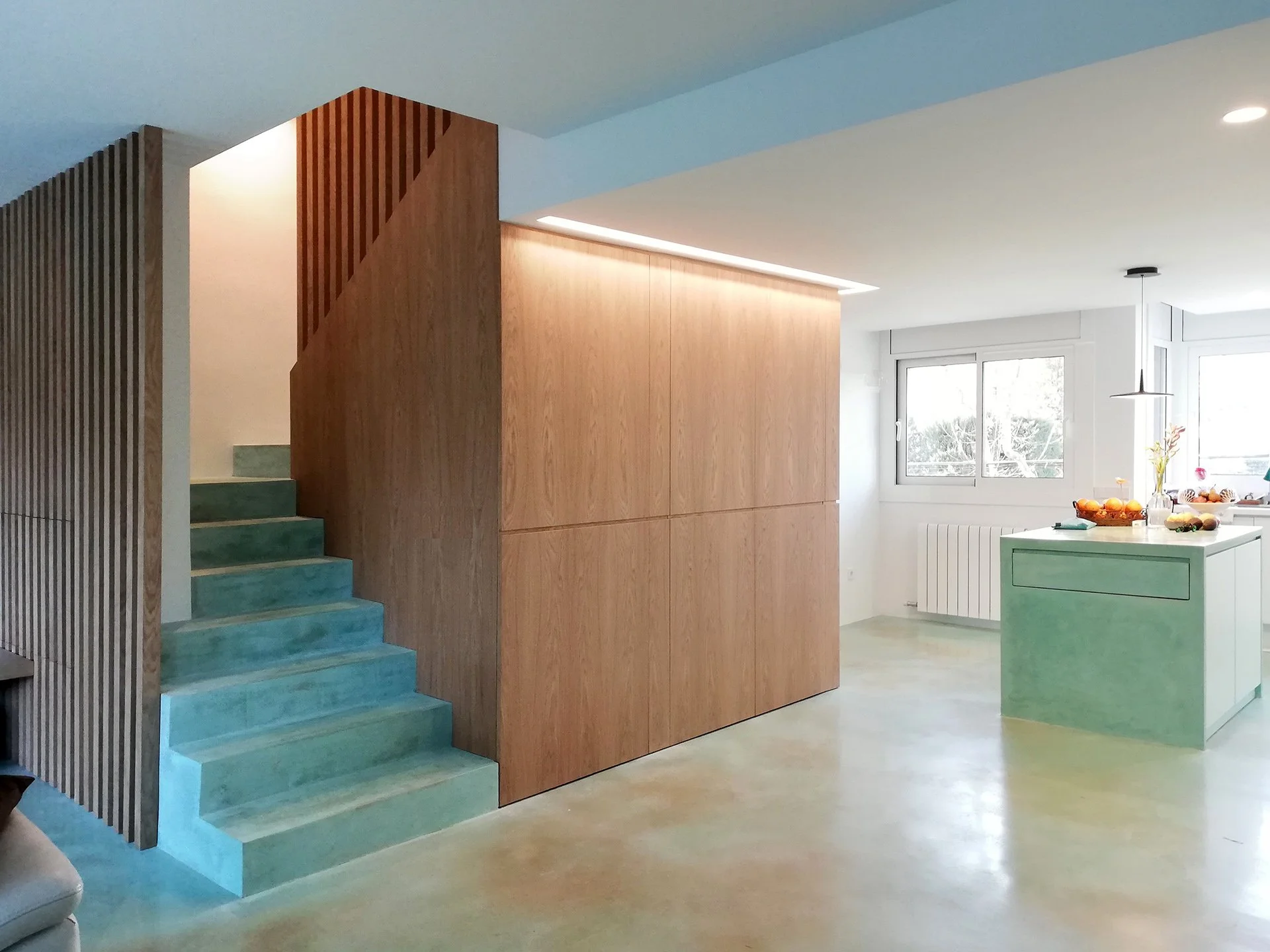 Espace intérieur moderne avec escalier en béton turquoise menant à un étage, cloison en bois clair, cuisine avec îlot vert pastel, fenêtres laissant entrer la lumière naturelle, décoration simple avec climat intérieur épuré.