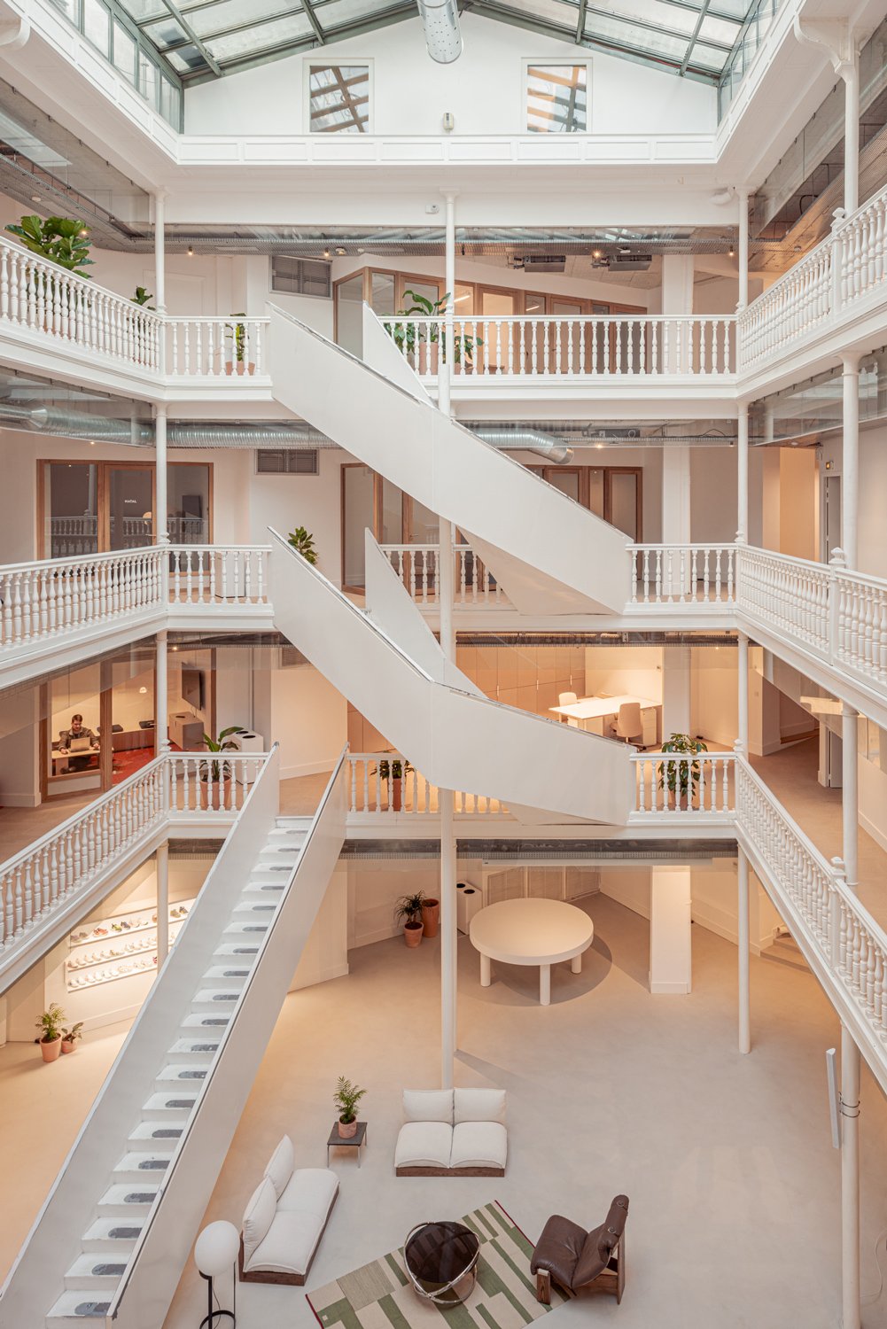 Intérieur moderne avec plusieurs étages, escaliers blancs en colimaçon, espaces de travail avec bureaux, coin détente avec sofa et fauteuil, plantes vertes, plafond en verre