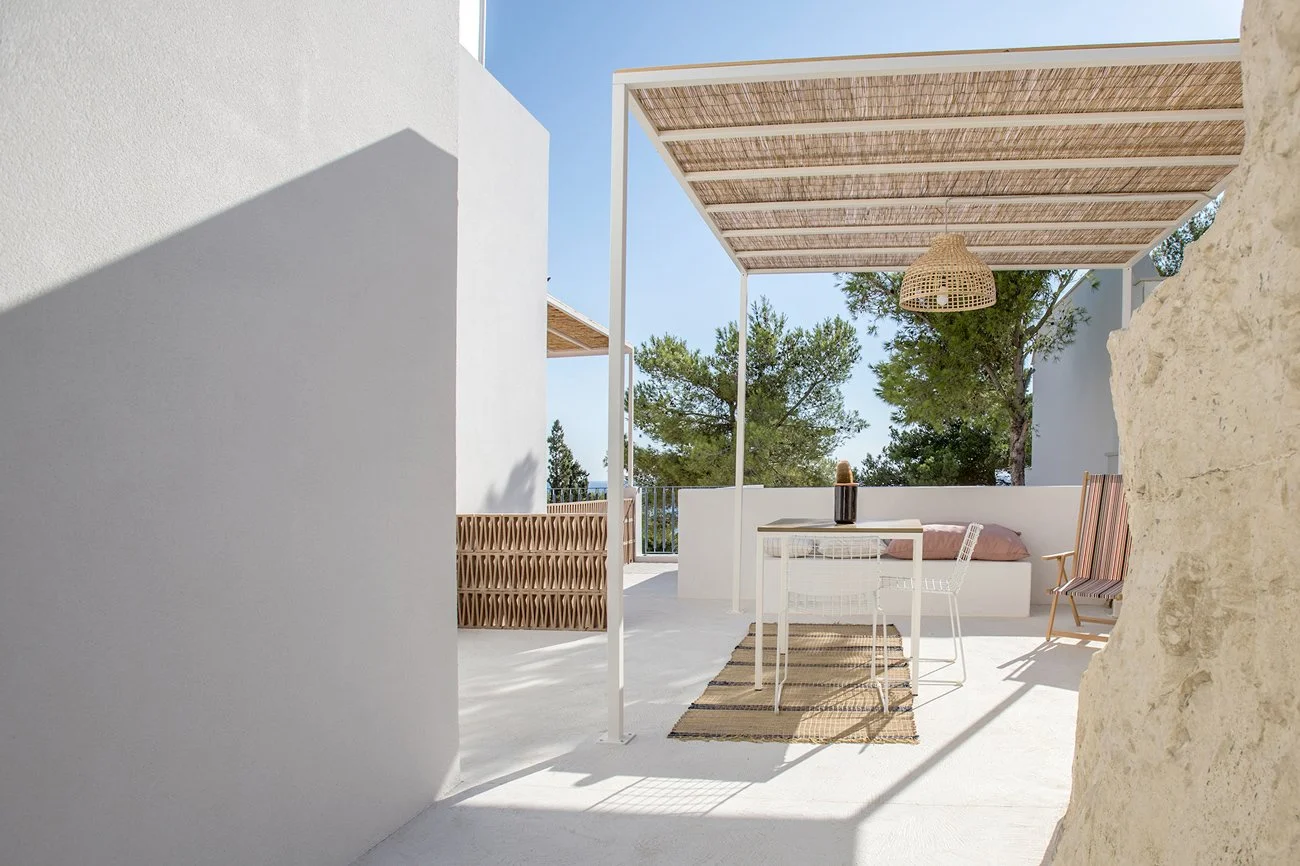 Terrasse ensoleillée avec mobilier en rotin et métal, pergola en bois, décor minimaliste, vue sur nature avec arbres, mur blanc et sol en béton blanc, ambiance méditerranéenne et moderne.
