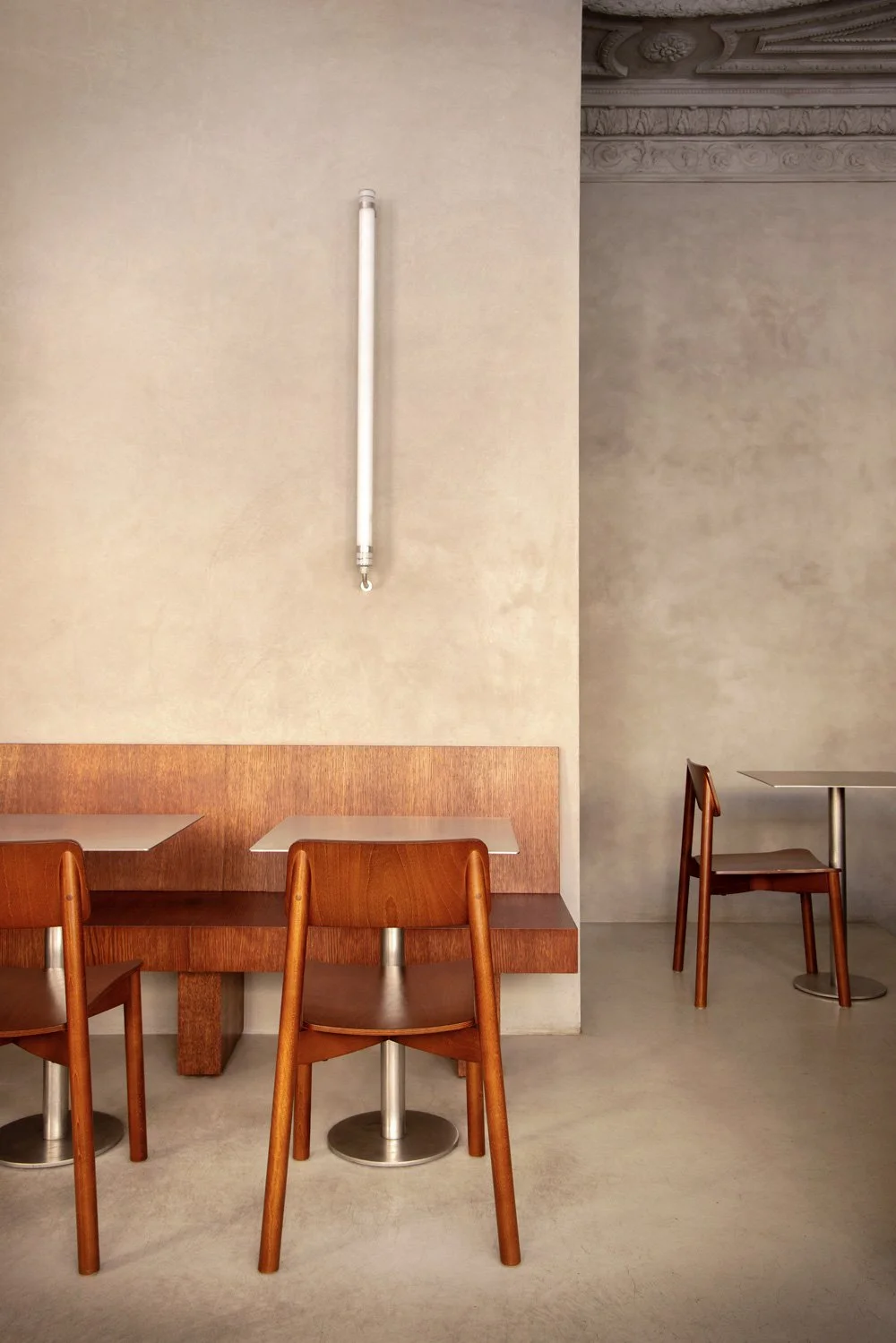 Intérieur minimaliste avec deux tables en bois et quatre chaises en bois, mur beige avec une barre lumineuse verticale, plafond décoratif en moulures.