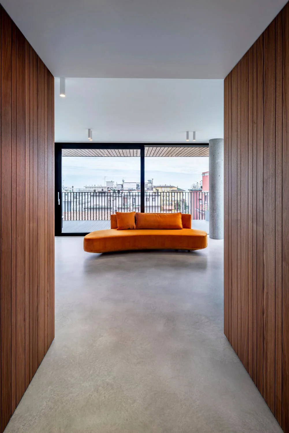 Un couloir avec des murs en bois menant à un salon moderne avec un canapé orange, une grande fenêtre donnant sur une vue urbaine et un sol en béton.