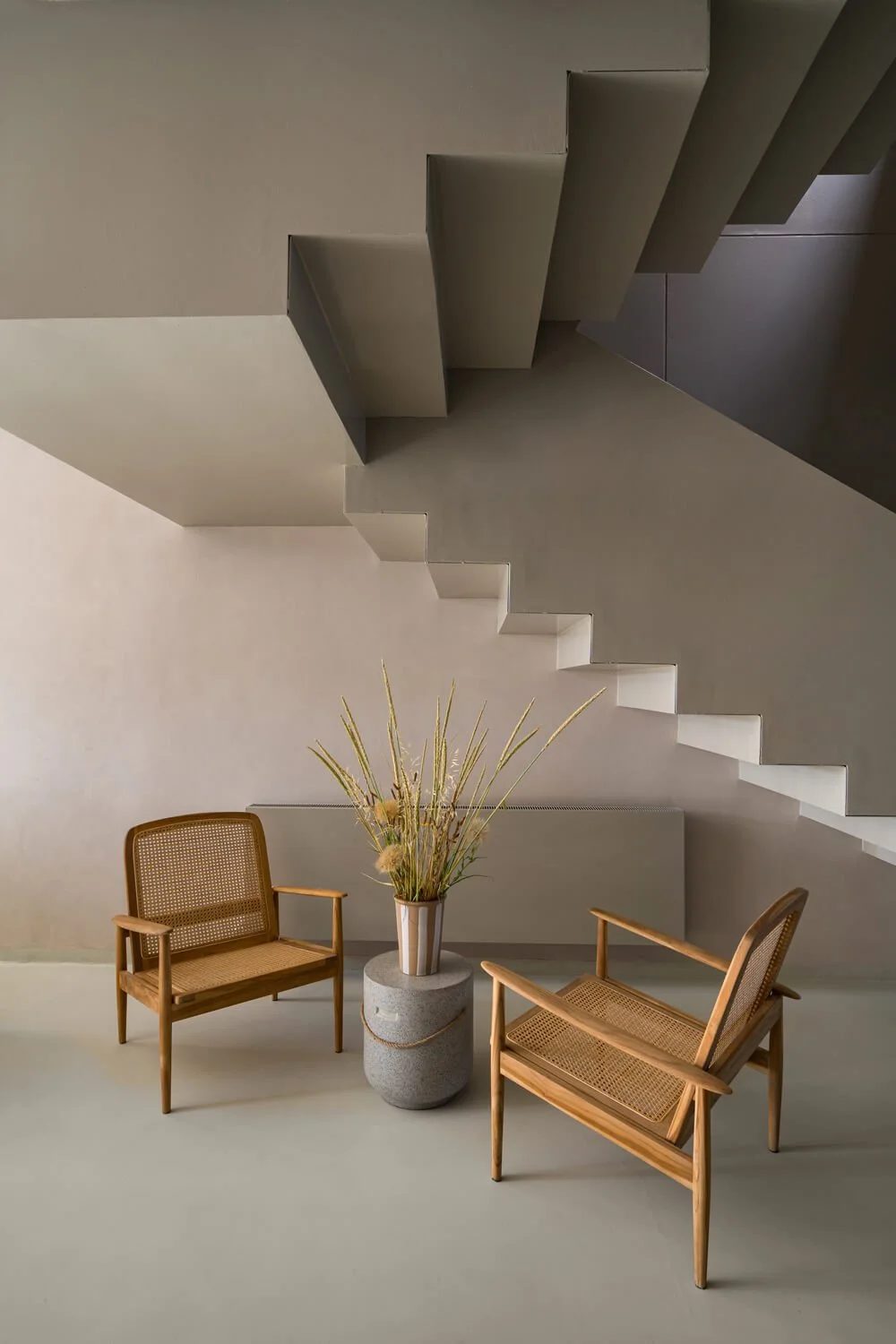 Deux fauteuils en bois avec cannage, une grande urne en pierre contenant des plantes sèche, sous un escalier moderne en gris avec des formes géométriques.