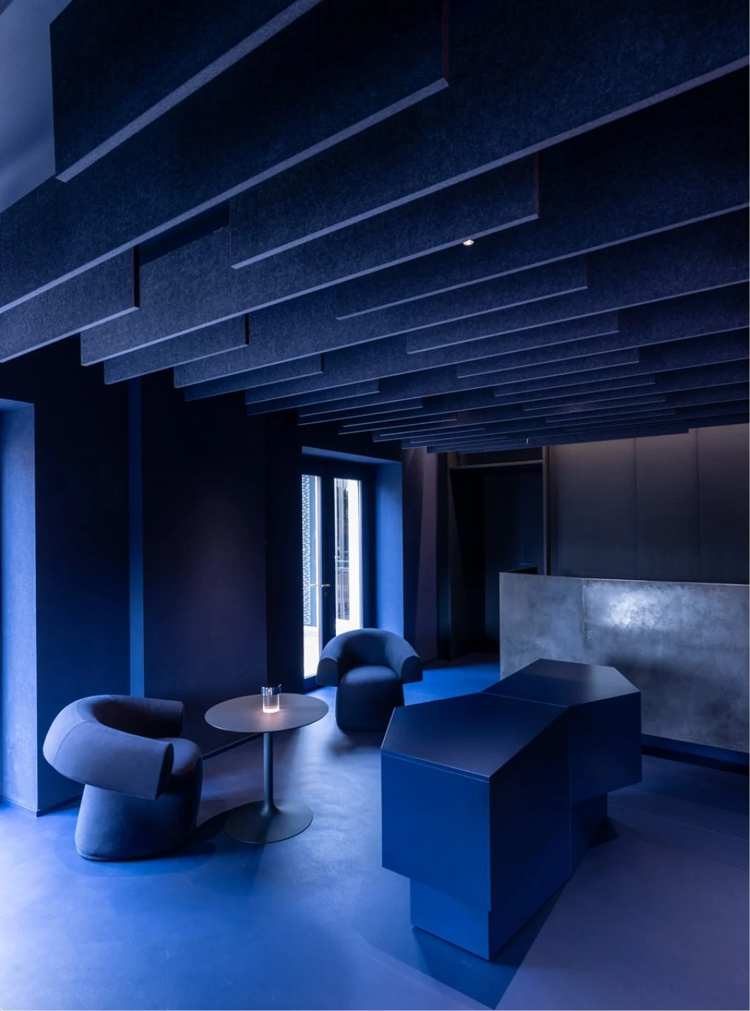 Intérieur sombre d'une pièce avec meubles modernes, comprenant deux fauteuils, une petite table ronde, un comptoir en béton, et des murs et plafond de couleur noire ou très foncée.
