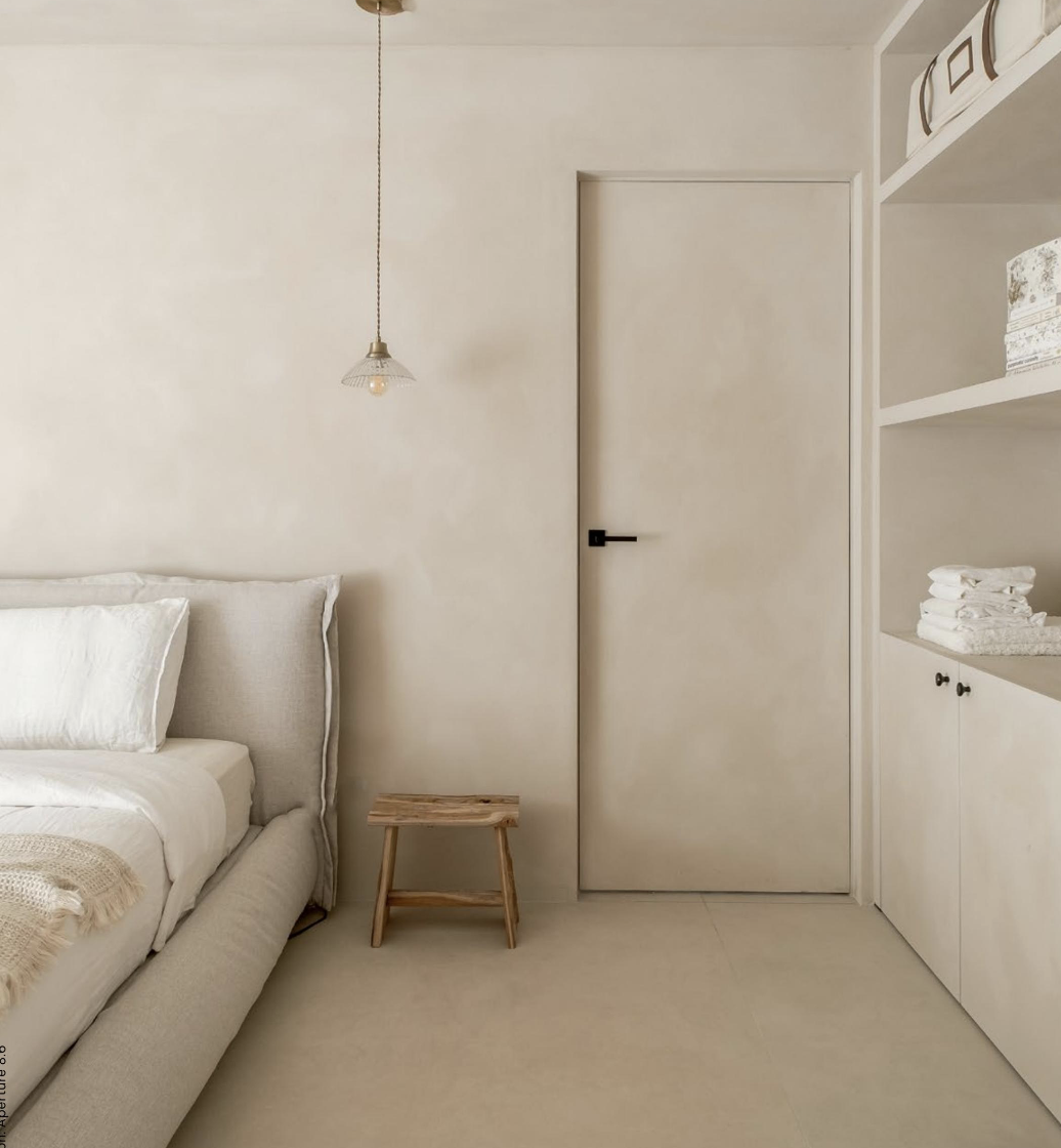 Chambre minimaliste avec un lit blanc, une petite table en bois, une porte blanche, et des étagères avec des serviettes et des boîtes de rangement.