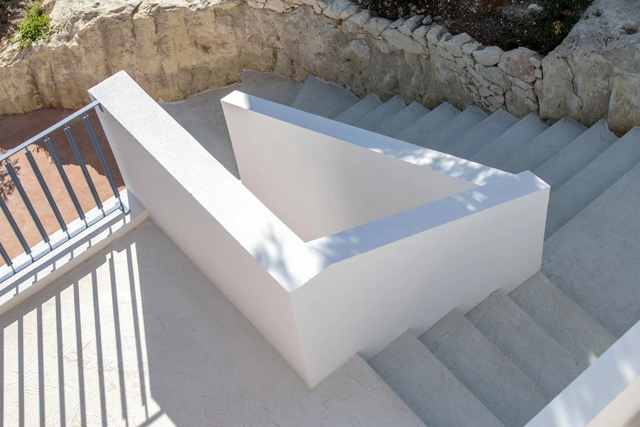 Escalier extérieur en béton blanc avec une barrière en métal, entouré de rochers et un mur en pierre.