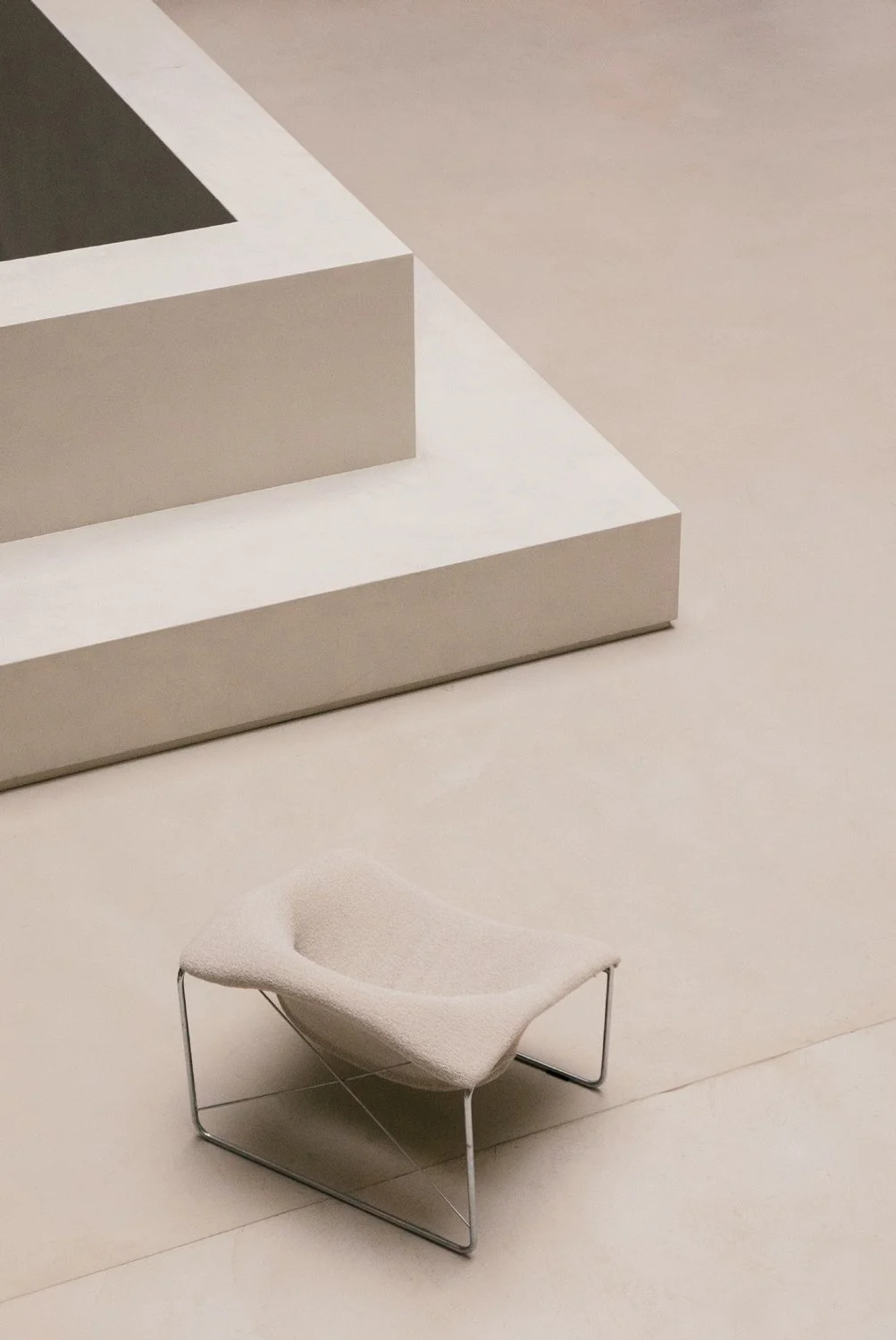 Une chaise beige avec une structure en métal au sol, dans une pièce avec des murs et un sol beige minimaliste.