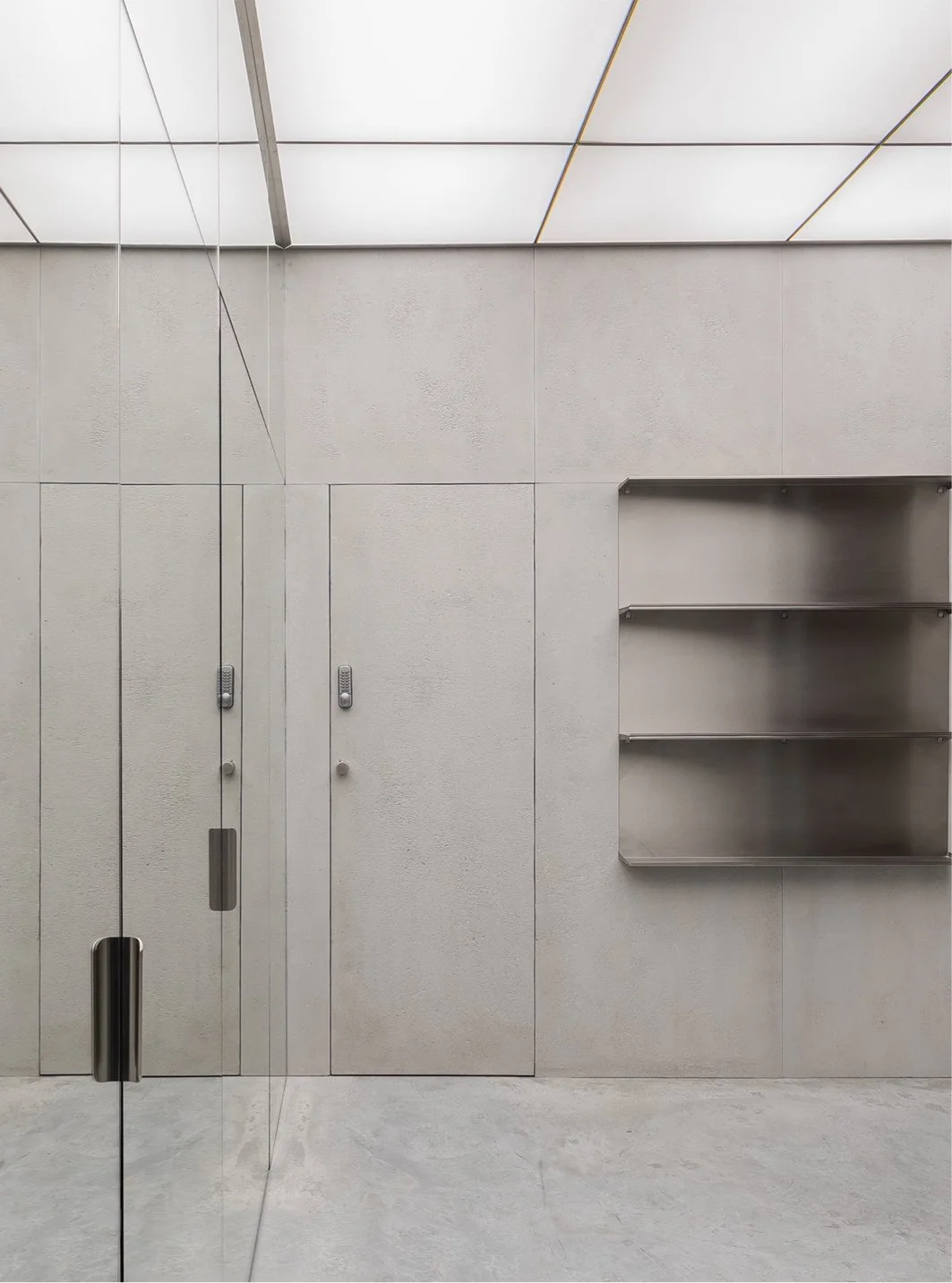 Photo d'une pièce minimaliste avec murs en béton clair, étagère en métal, porte en verre côté gauche avec poignée en métal, et plafond en panneaux lumineux.