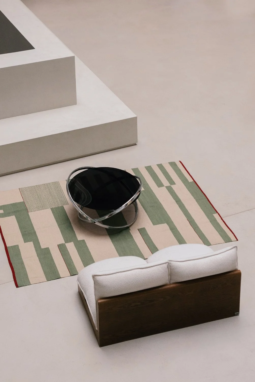 Une scène intérieure avec un tapis à motifs géométriques verts et beiges, un bol noir avec un cadre métallique, une banquette en bois avec coussin blanc, et des marches blanches.