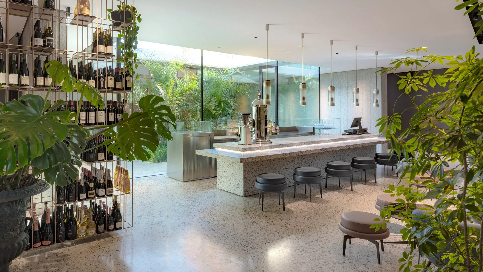 Intérieur moderne d’un bar avec un long comptoir en pierre, des tabourets noirs, des étagères avec bouteilles de vin, et des plantes vertes en décoration.