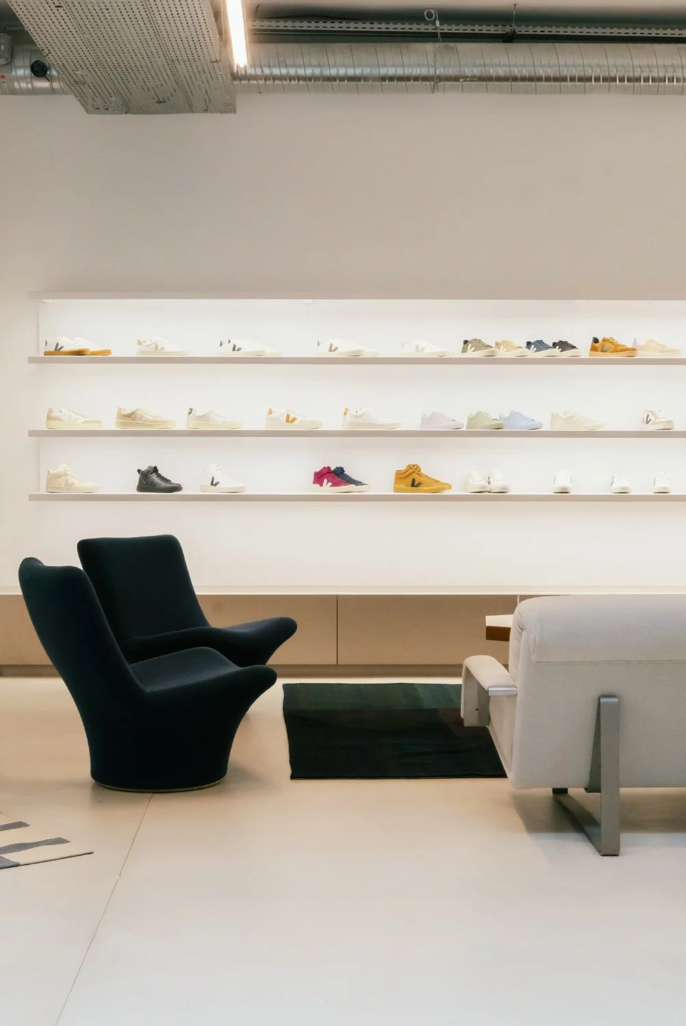 Étagère éclairée présentant une collection de sneakers colorés dans une boutique, avec deux fauteuils noirs et un canapé blanc devant eux.