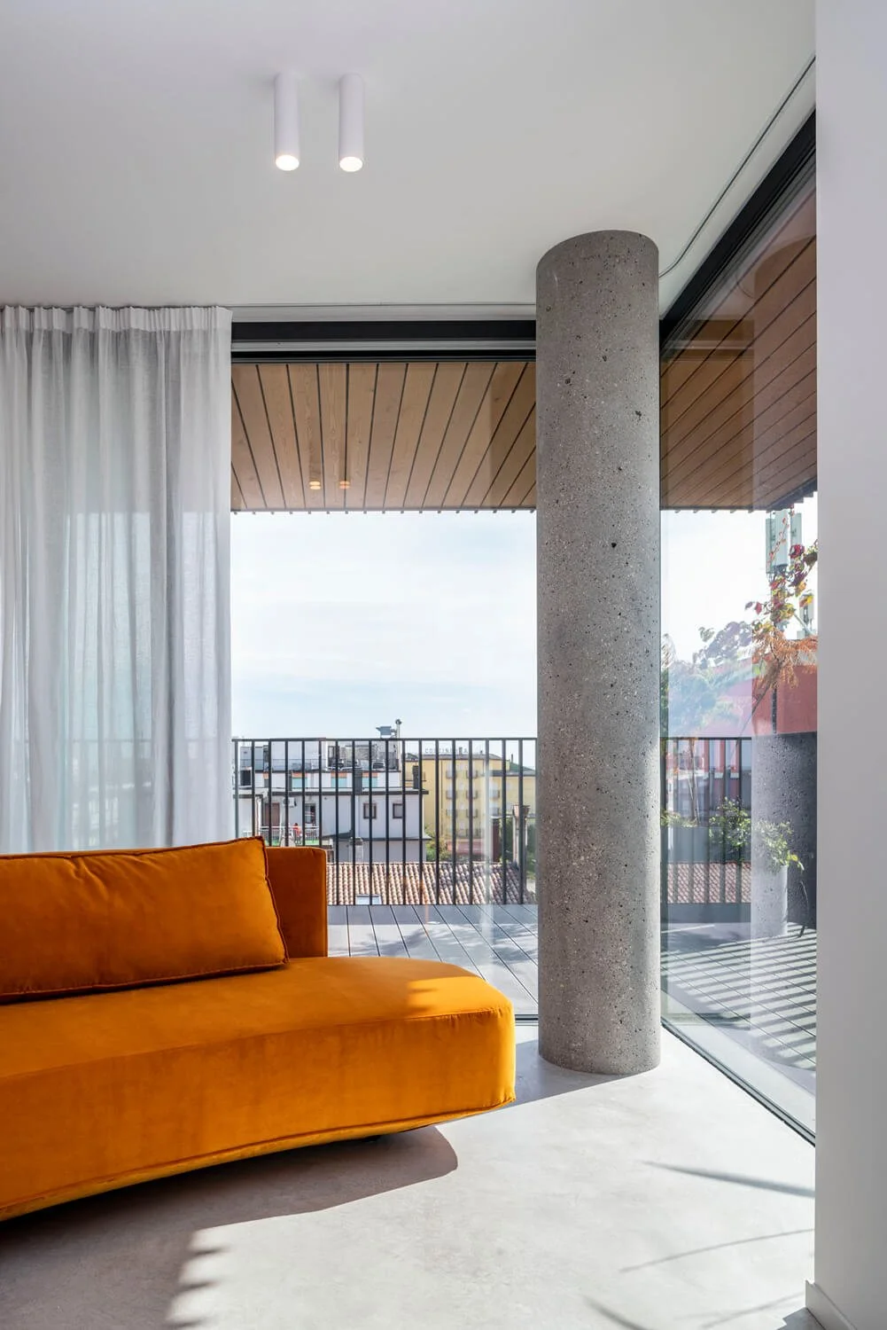 Salon moderne avec un canapé orange et une vue sur une terrasse avec un plafond en bois, un pilier en béton et de grandes fenêtres.
