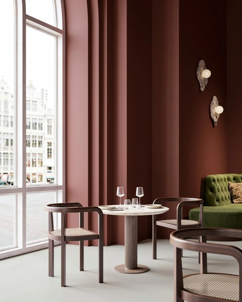 Intérieur d'un salon avec un mur rose, grande fenêtre, table ronde avec trois verres à vin, fauteuils en bois et un canapé vert avec coussin décoratif.