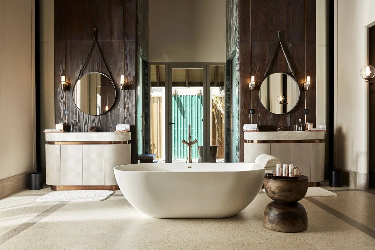 Salle de bain avec une baignoire blanche centrale, deux plans de lavabo avec miroirs ronds, décoration élégante en bois et en beige, lumière naturelle