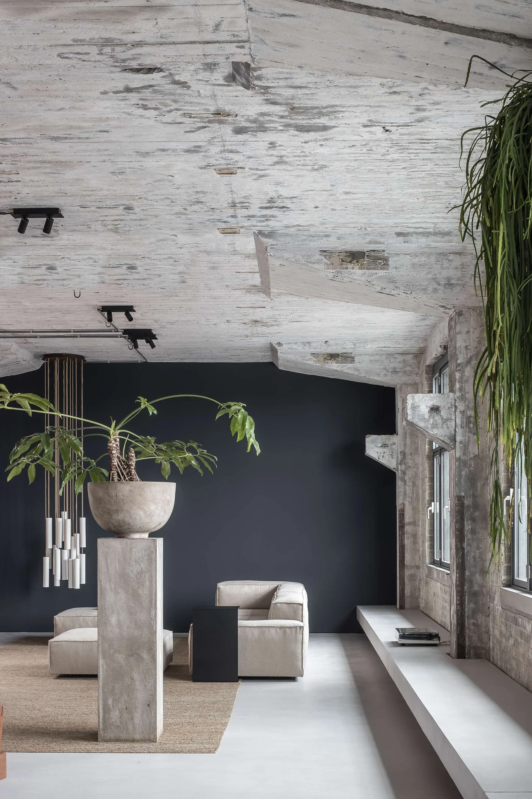 Intérieur moderne avec murs en briques apparentes et plafond en bois blanc, comprenant un canapé beige, une plante en pot sur un socle en béton, une étagère intégrée en blanc et des fenêtres laissant entrer la lumière naturelle.