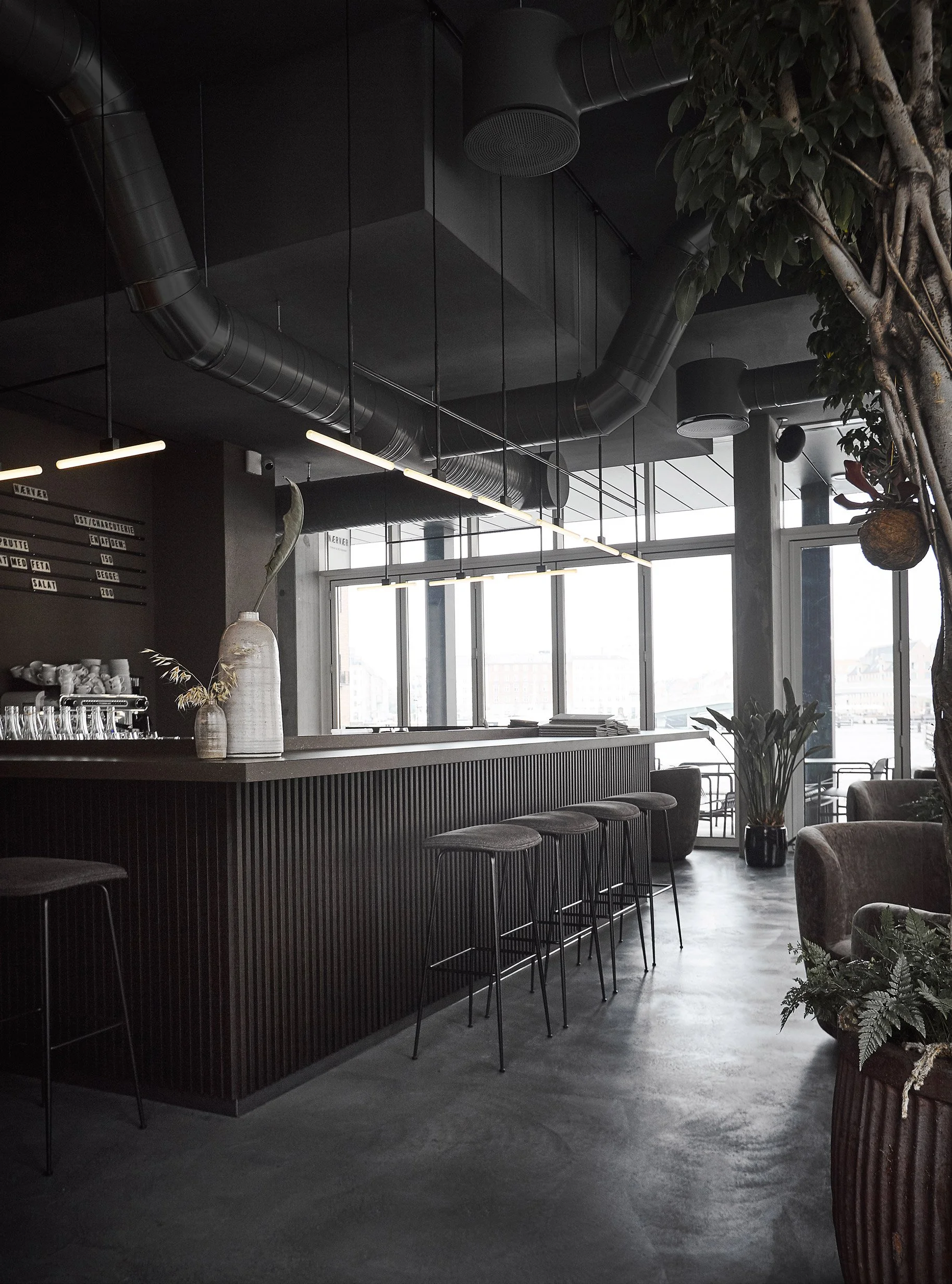 Intérieur d'un café ou bar moderne avec comptoir en bois sombre, tabourets, grande fenêtre laissant entrer la lumière naturelle, plantes d'intérieur et plafonds avec conduits d'air noirs.