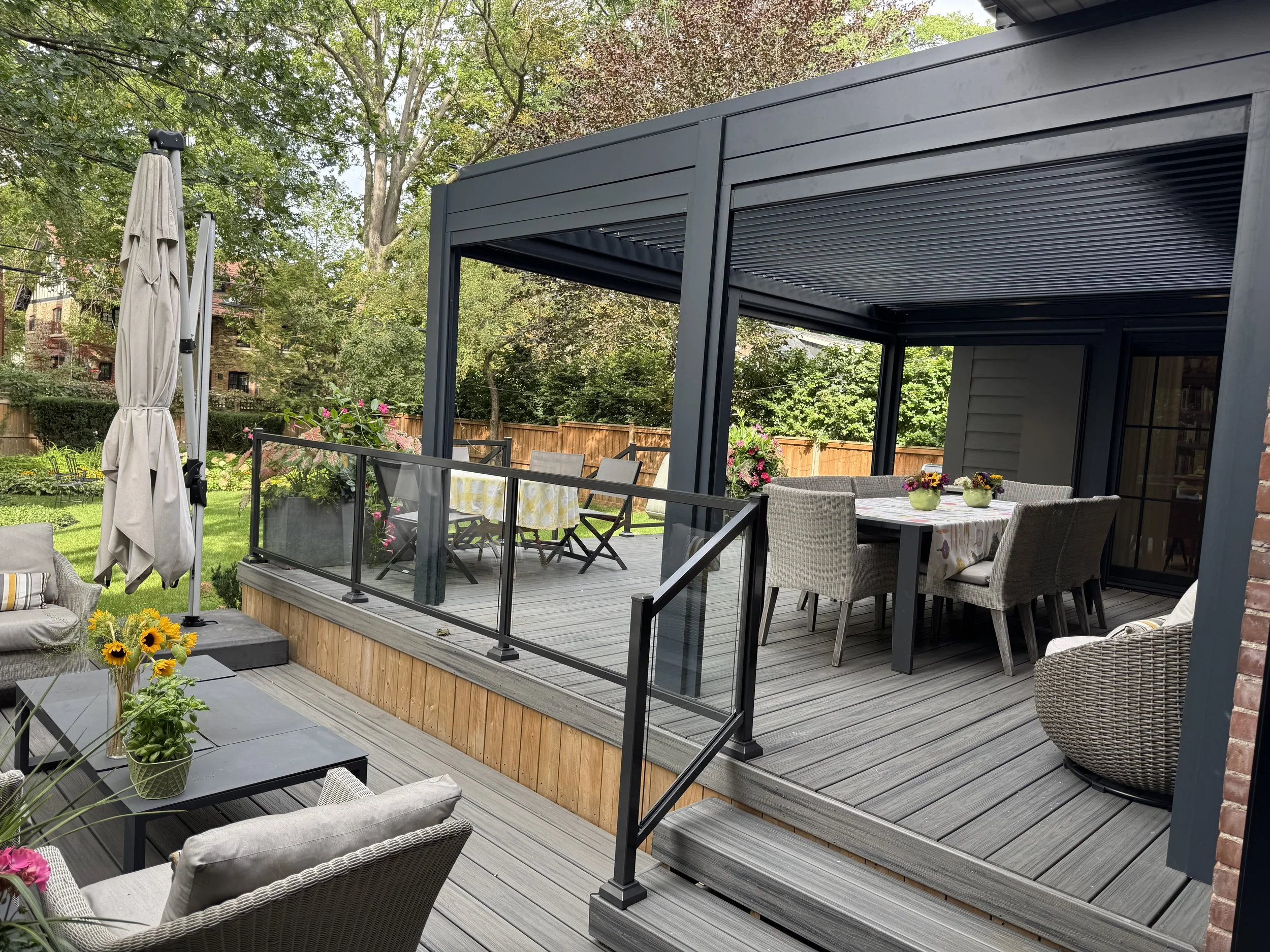 A backyard patio with a modern dark gray pergola, outdoor dining table with chairs, potted flowers, and a covered sun umbrella, surrounded by trees and a wooden fence.