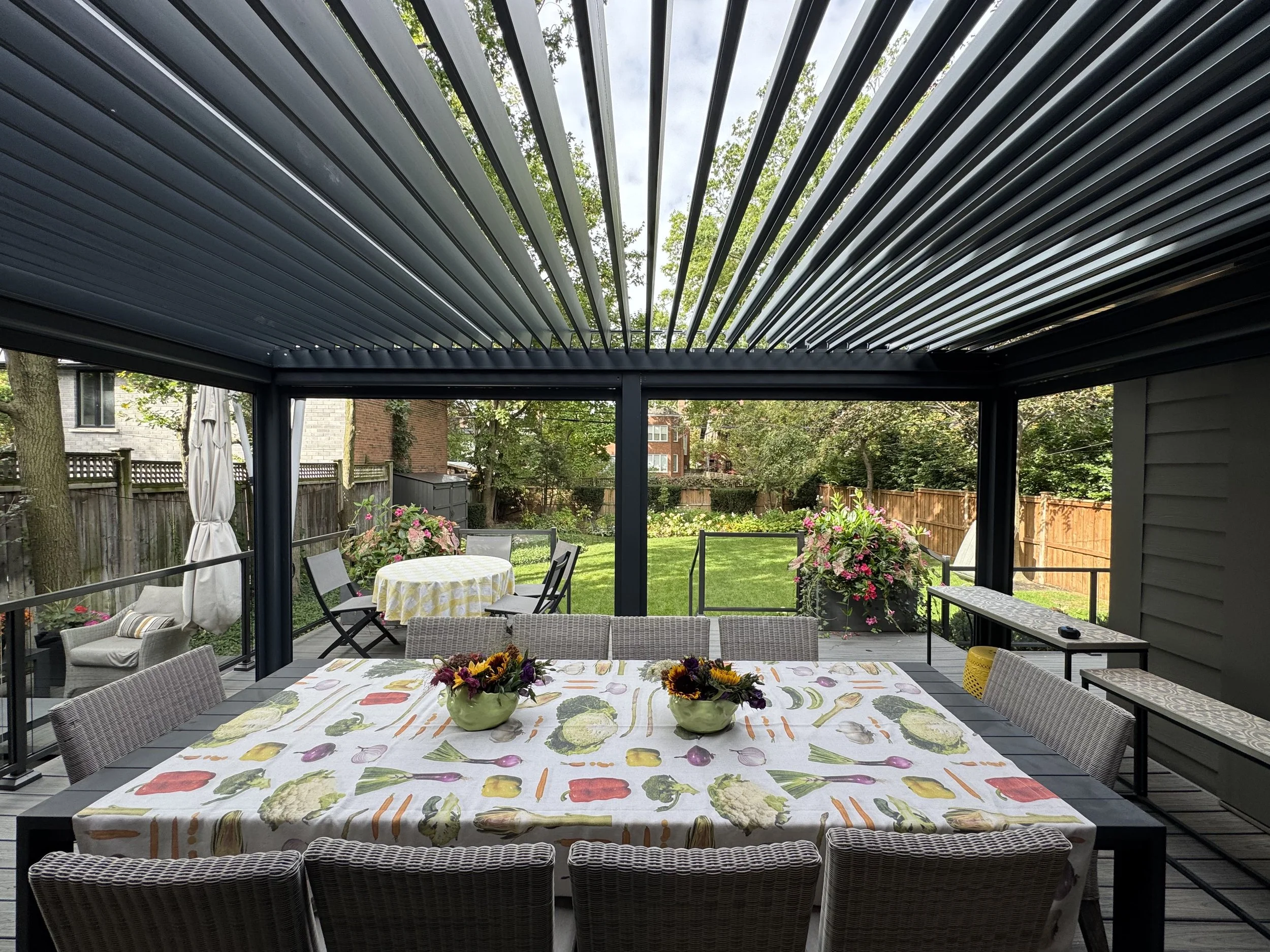 An outdoor patio area with a dining table covered with a colorful vegetable pattern tablecloth, decorated with two green vases filled with flowers. The patio has a table, chairs, and tropical plants, with a surrounding wooden fence and trees in the b