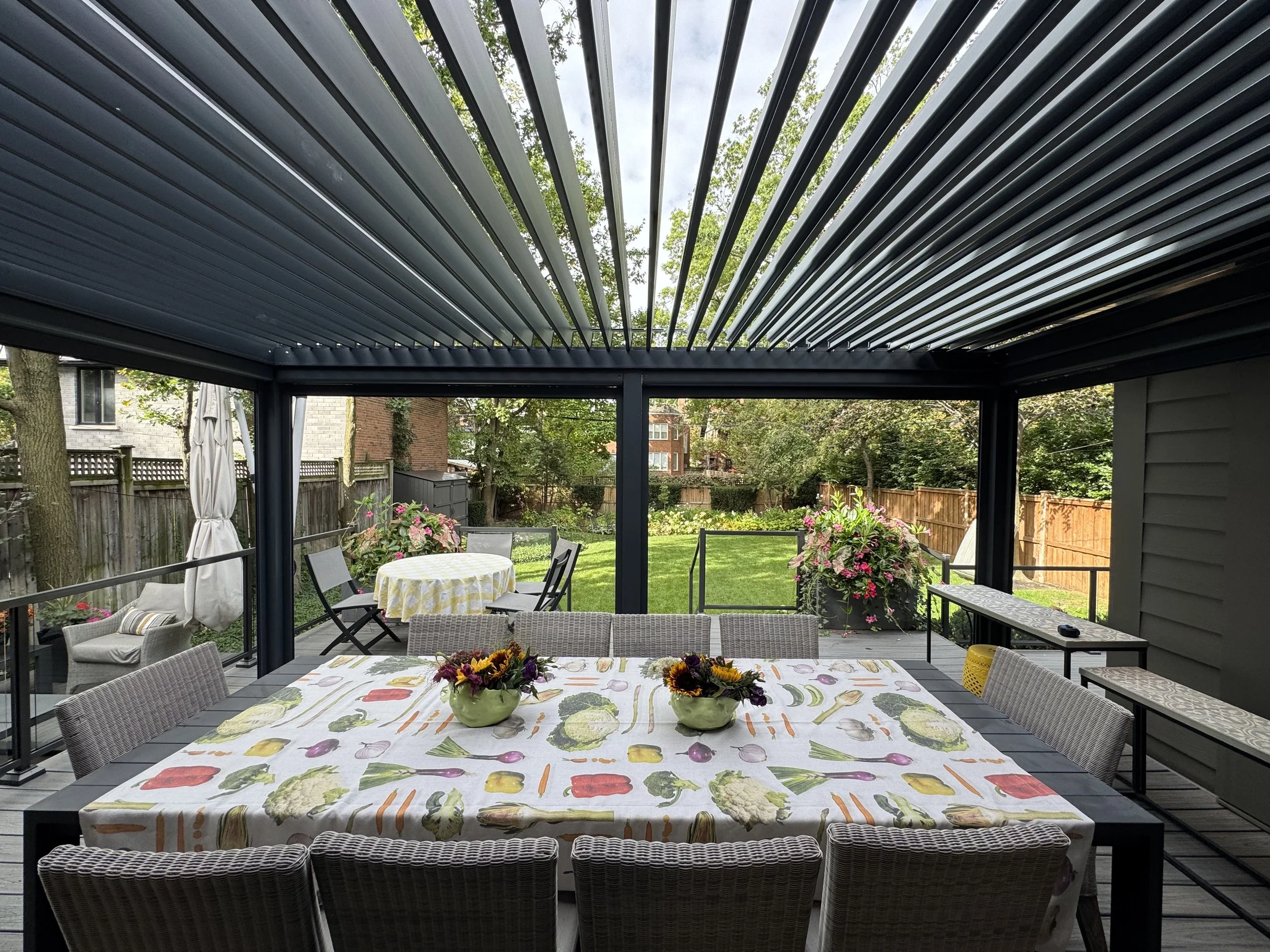 A covered outdoor patio with a table covered with a vegetable-themed tablecloth, decorated with centerpieces of sunflowers and other flowers, surrounded by chairs, with a garden and houses in the background.