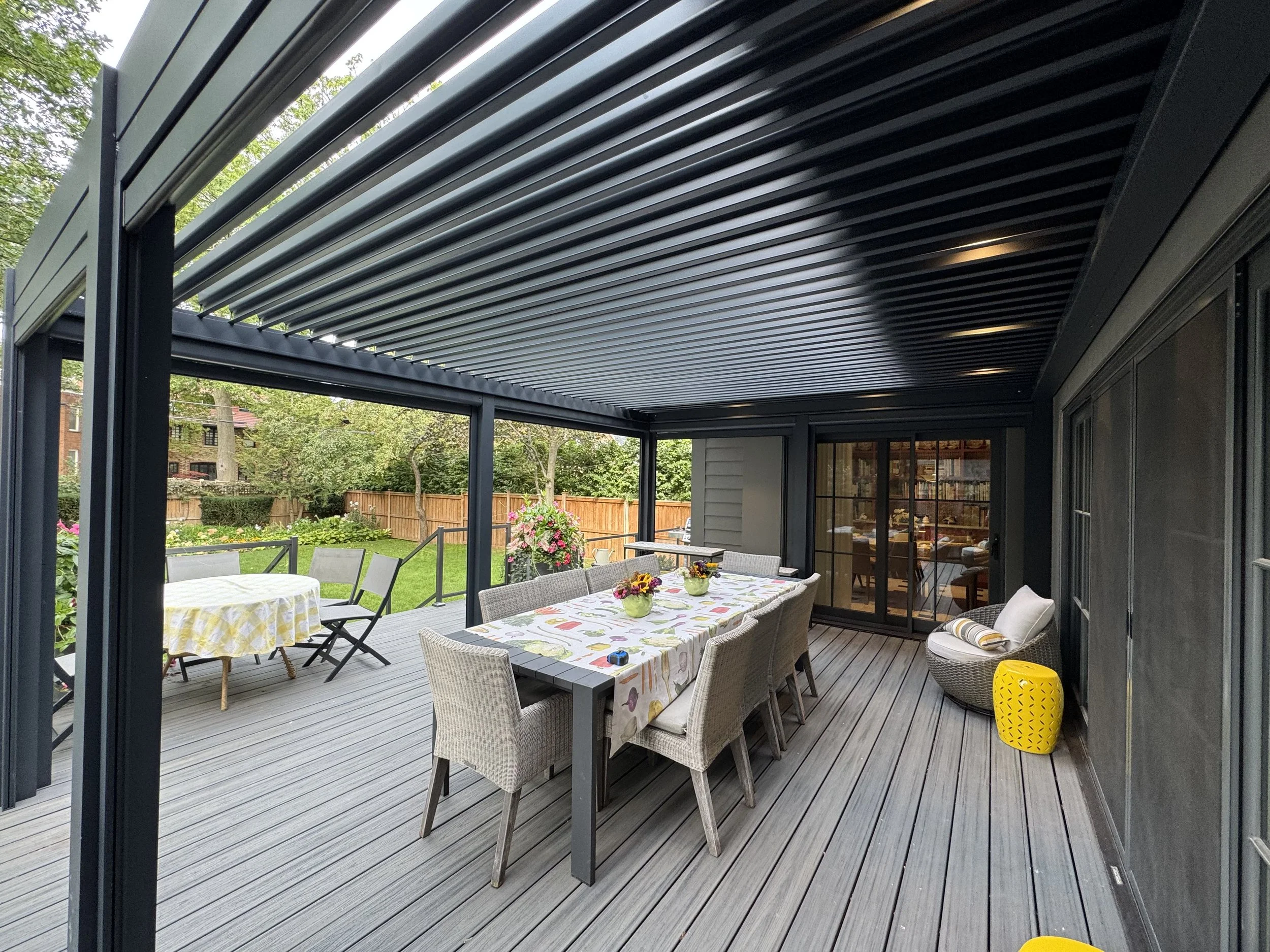 A covered outdoor patio with a dining table, chairs, potted plants, and a view of a backyard with green grass, trees, and a wooden fence.