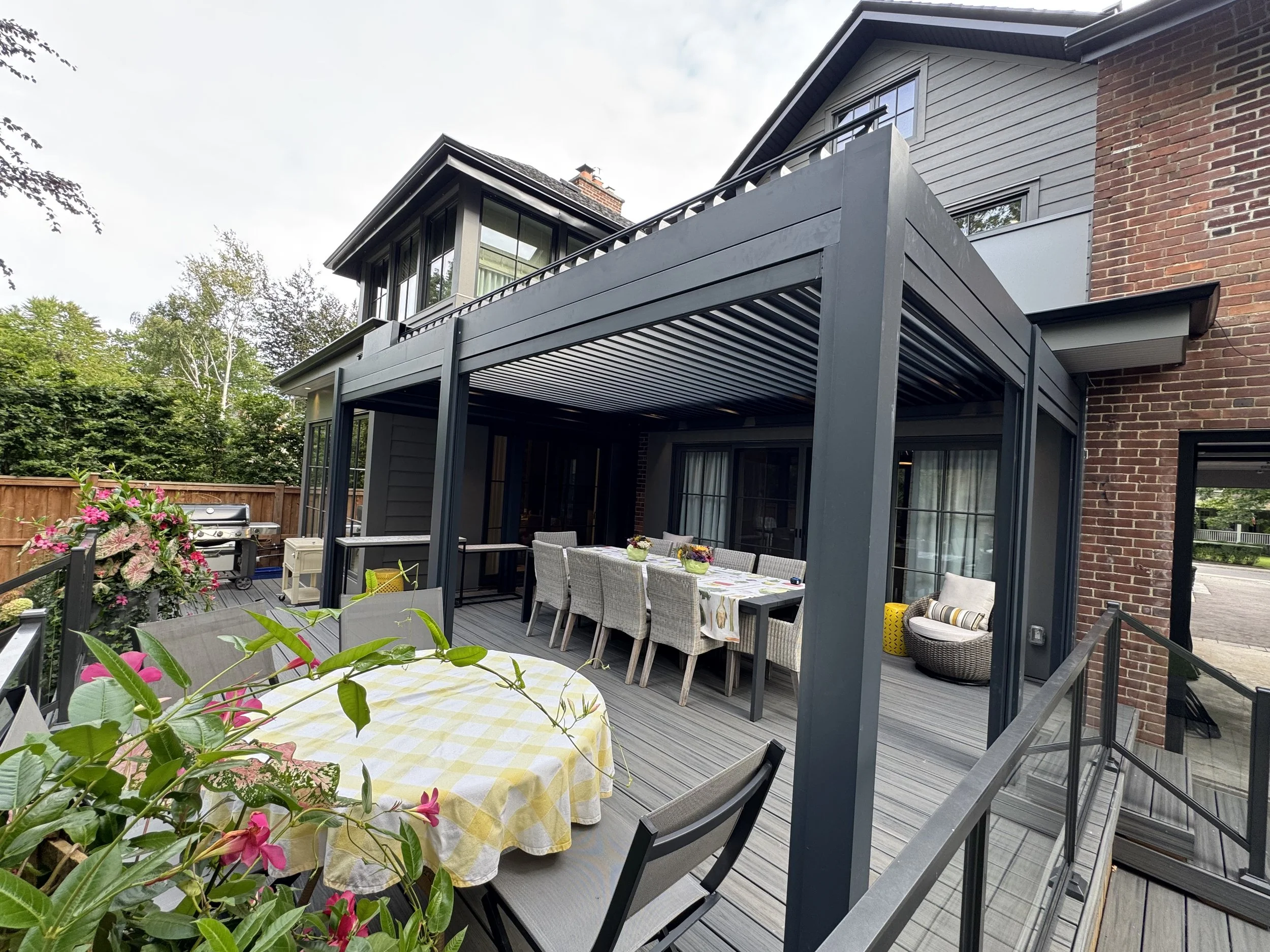 Backyard patio with dining table covered with a yellow checkered tablecloth, surrounded by chairs, under a pergola, with potted plants and flowers, adjacent to a brick house with large windows