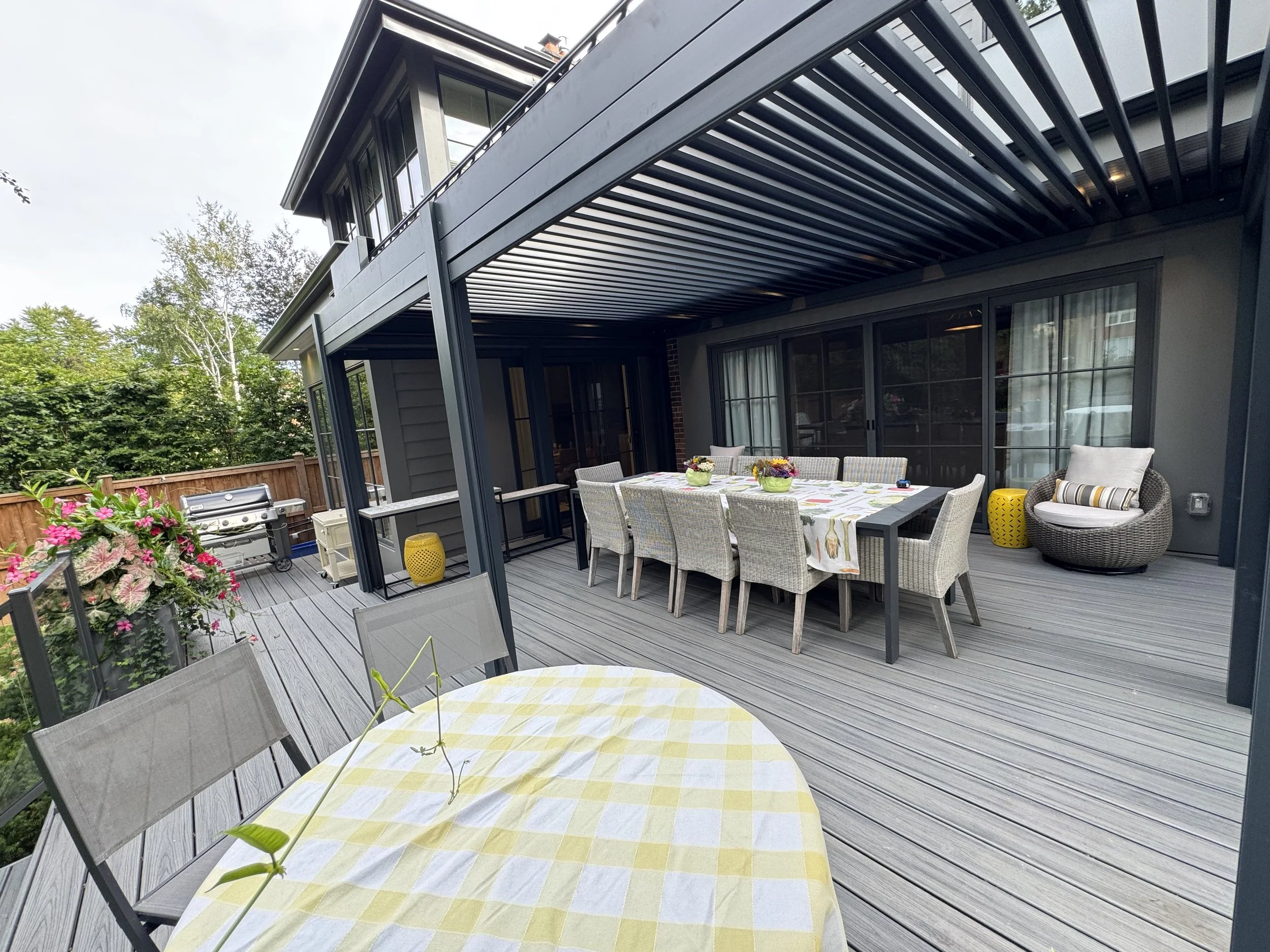 A backyard patio with a dining table covered with a yellow and white checkered tablecloth, surrounded by chairs, and decorated with flower arrangements. There is a grill on the left, and the patio is shaded by a modern, black pergola. The background 