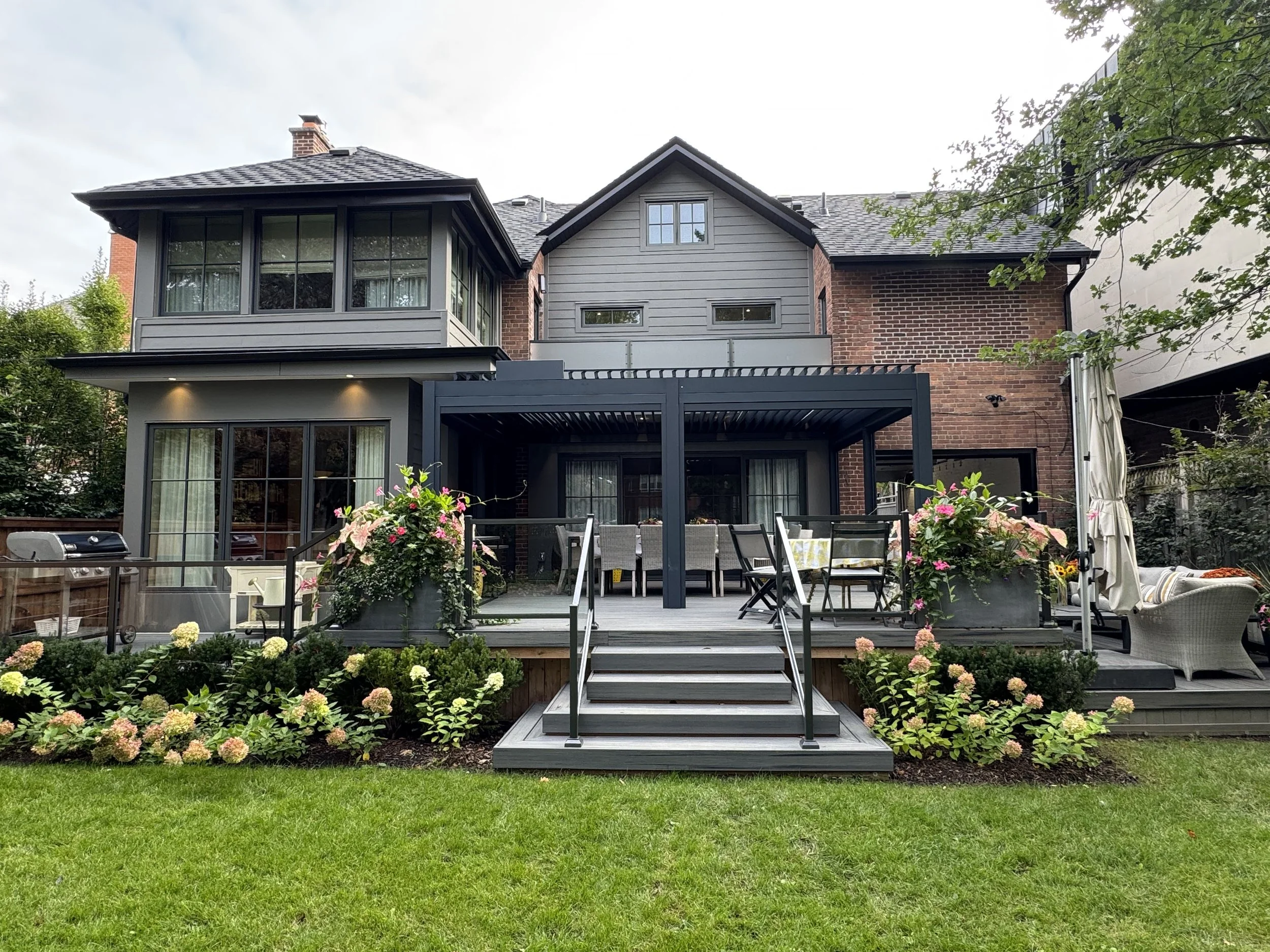Backyard with a raised deck, outdoor dining table with chairs, pink flowers in planters, garden with hydrangeas, green grass, and a multi-story house with brick and siding exterior.