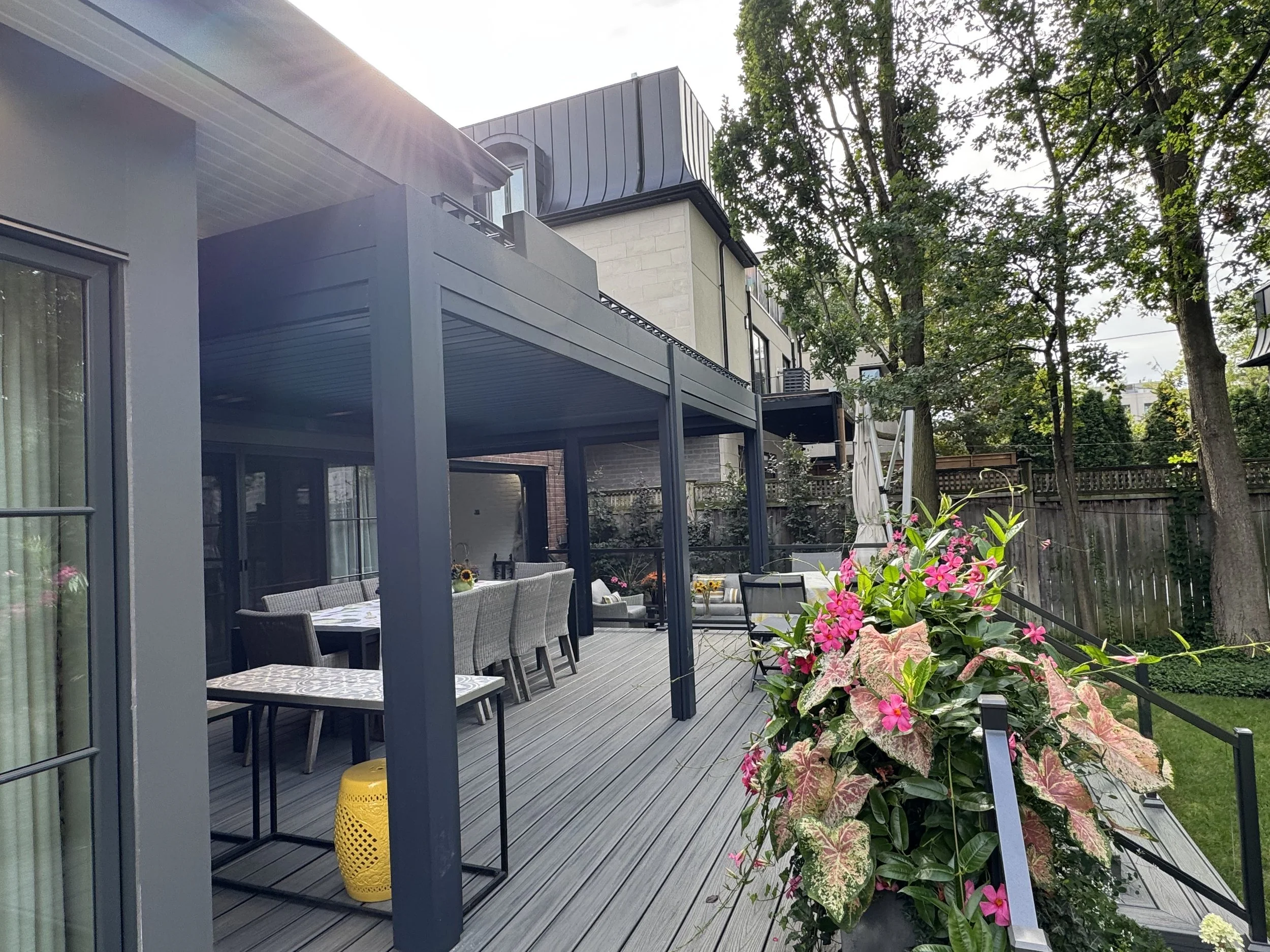 View of a backyard patio with outdoor dining table and chairs, potted pink flowers, and a house with large windows and a balcony, surrounded by trees and a wooden fence, during daytime.