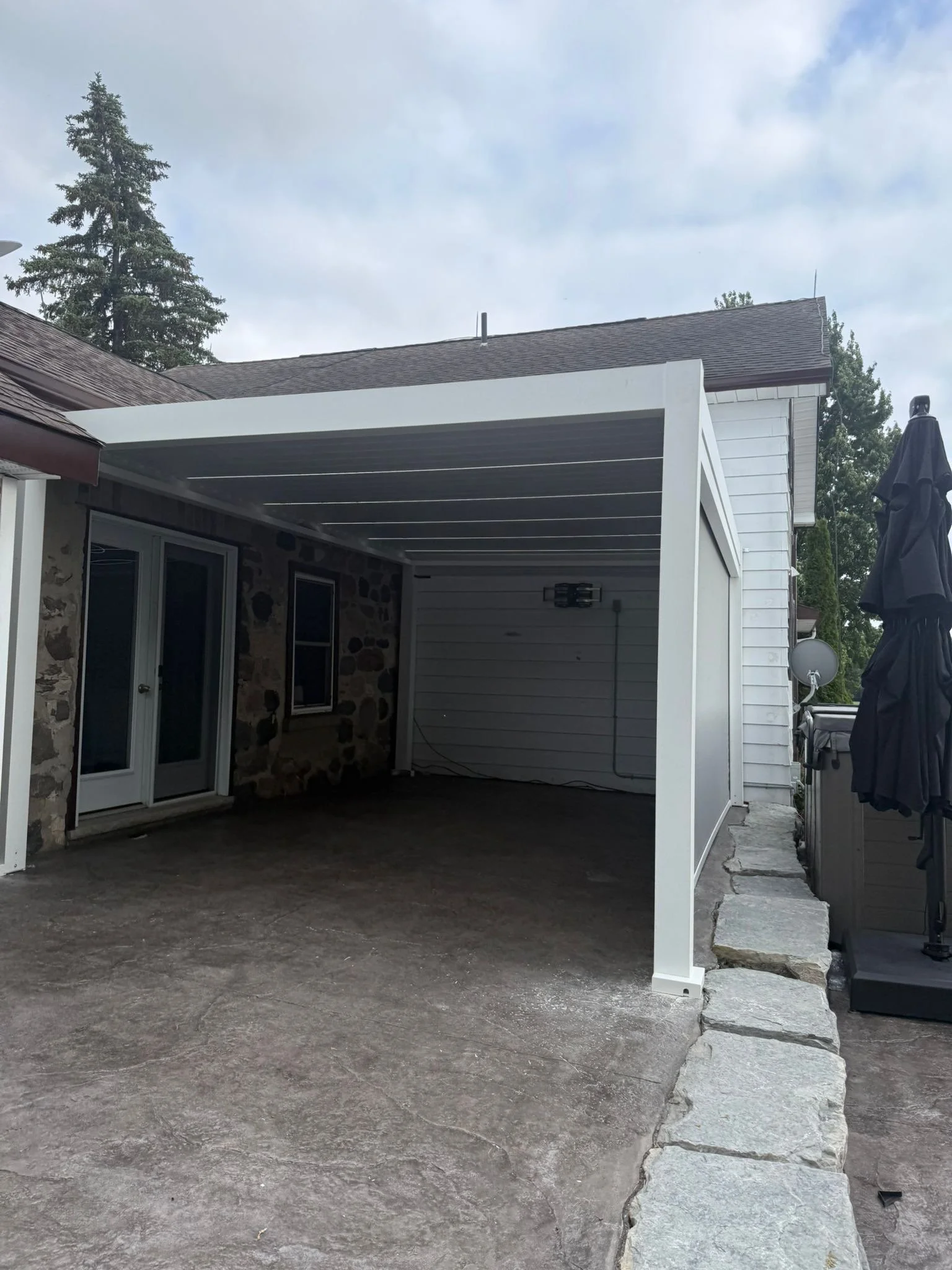 A covered outdoor patio area attached to a house, with a concrete floor, right side featuring a grill, and a white horizontal panel above. There are large rocks lining the edge of the patio and a view of a cloudy sky with trees in the background.