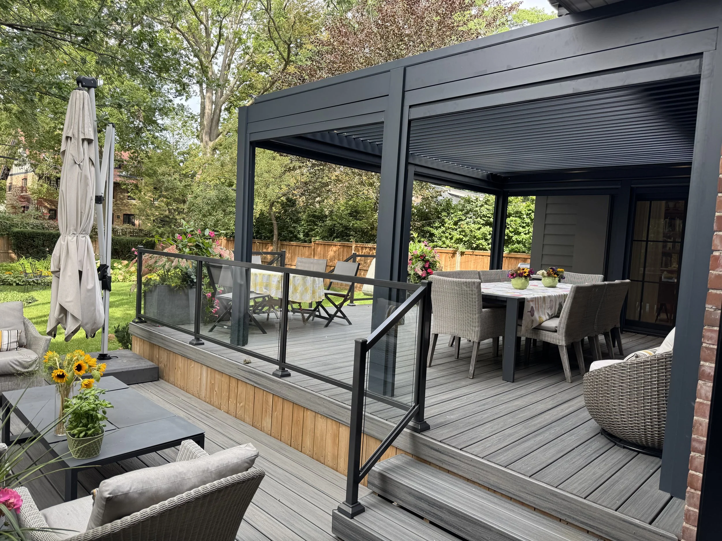An outdoor patio with a dining table, chairs, and potted flowers, extending from a house with a covered porch, surrounded by greenery and trees.