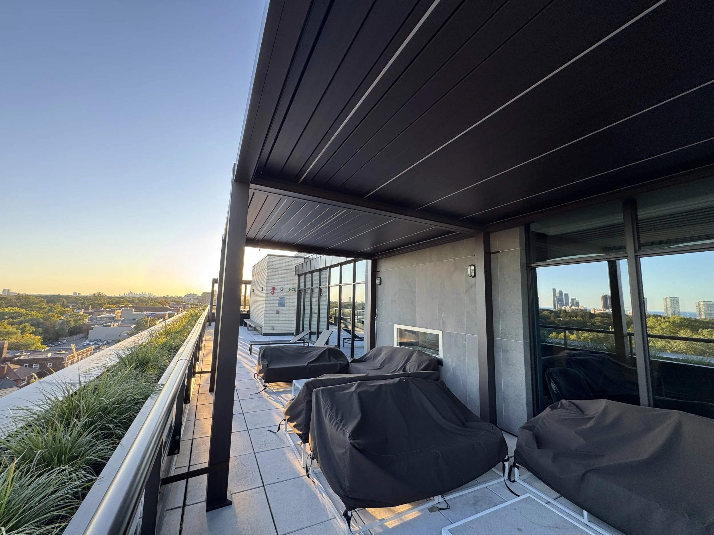 View from a balcony showing black seating and a roof with thin white lines, overlooking a city skyline and greenery during sunset.